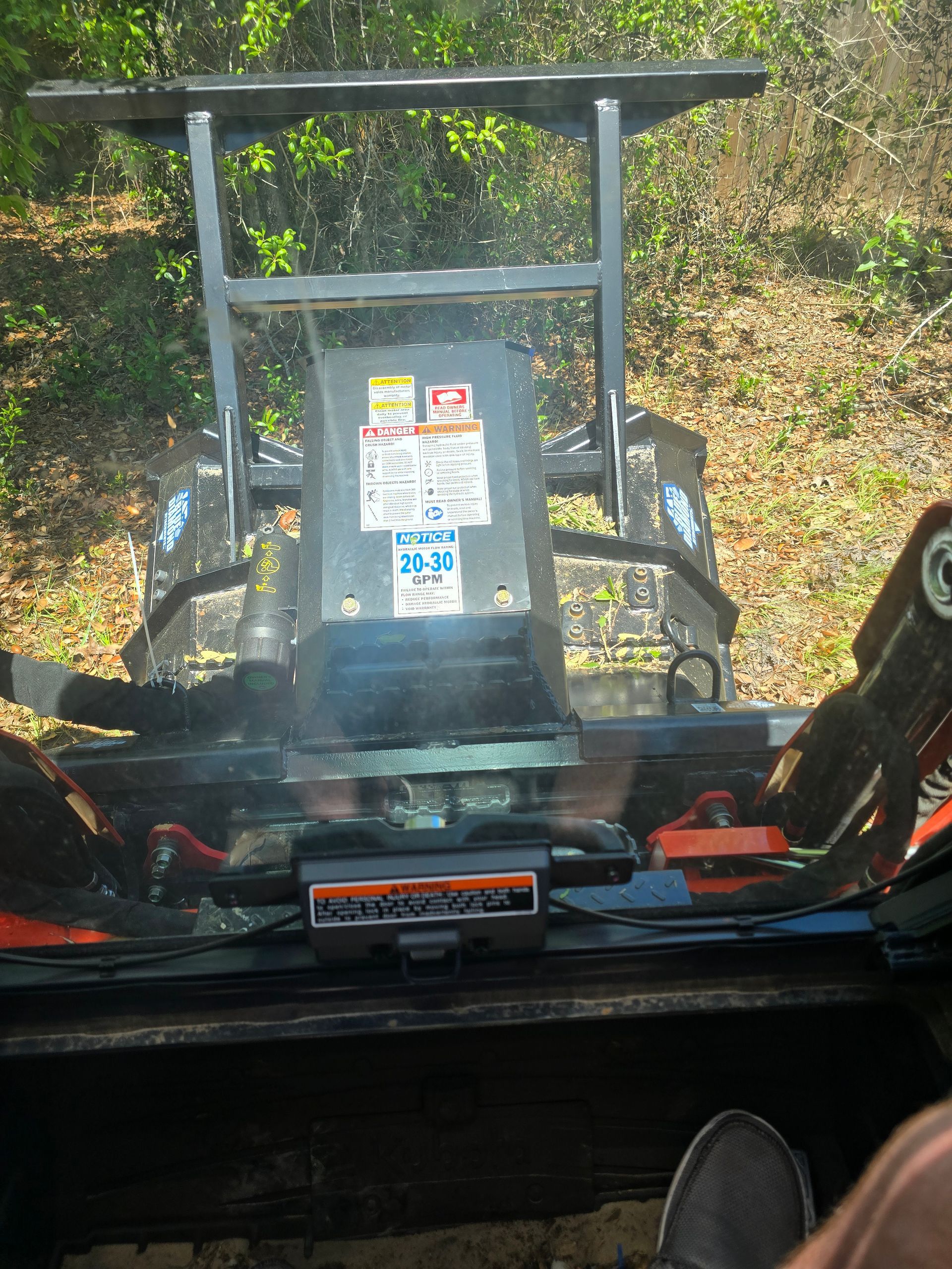 View from the cab of an orange skid steer, looking forward through the windshield at a metal brush cutter attachment.