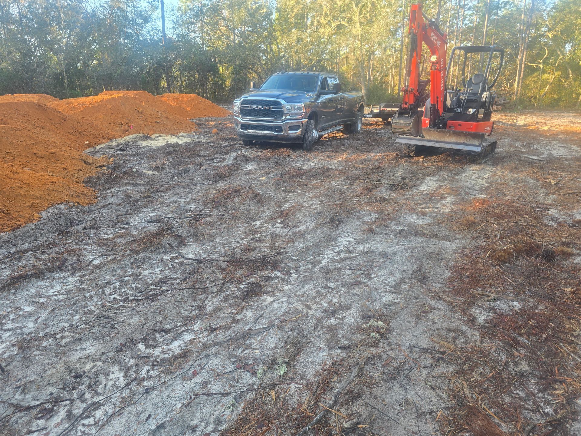 A gray pickup truck and an orange excavator parked on a clearing of dirt and wood chips with piles of soil nearby.