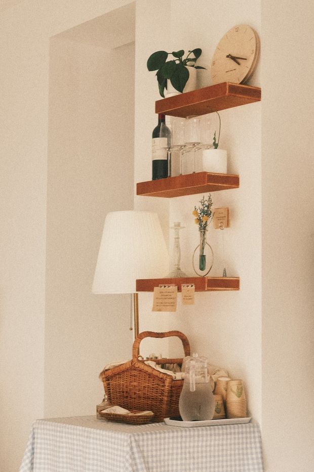 Shelves with decor: plant, wine, clock, vase. Table with basket, lamp, drink, and food.