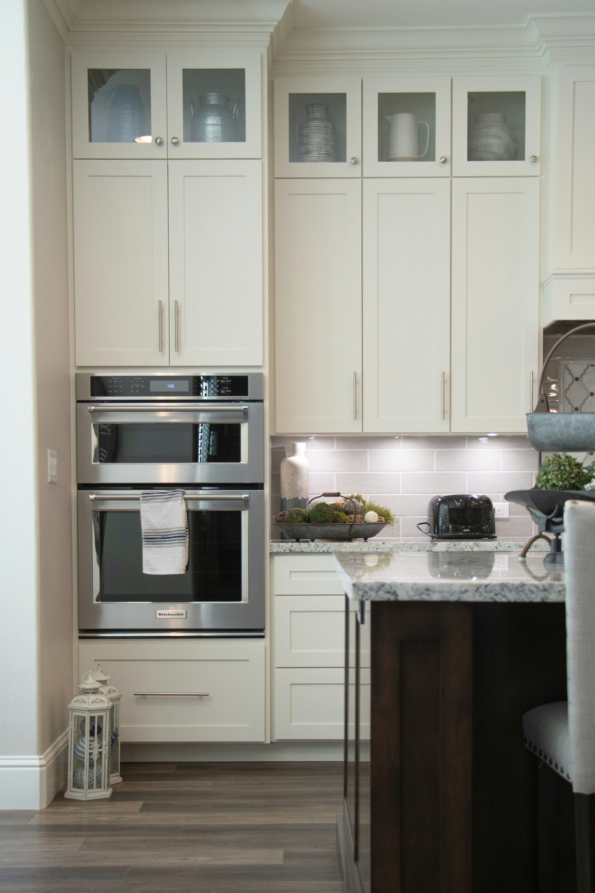 White kitchen cabinets with built-in stainless steel double oven, counter with items, and dark wood island.