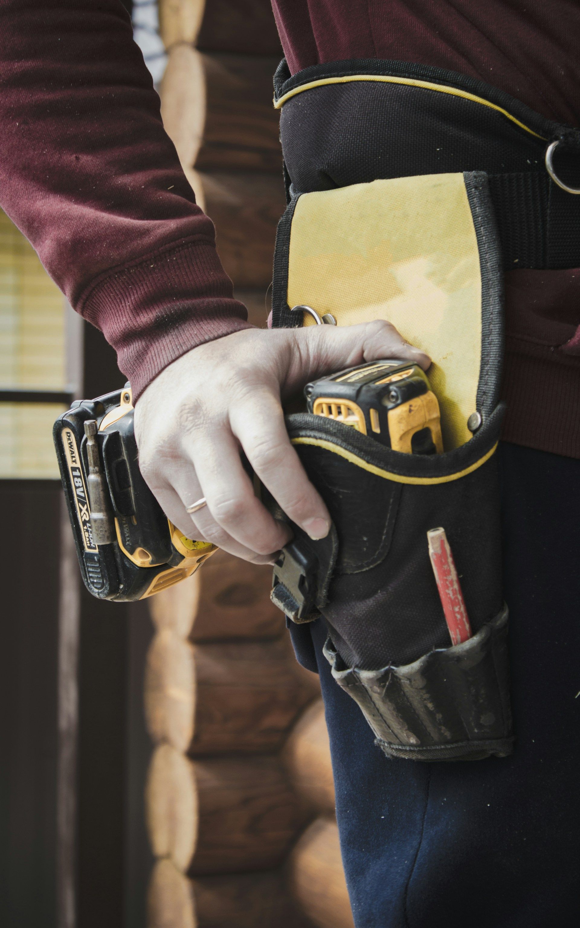 Person wearing a black and yellow tool belt, holding a drill. Log cabin background.
