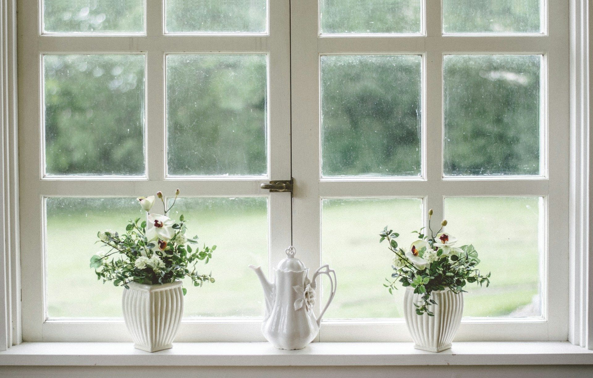White window with teapot and vases of flowers on the sill, looking out to a green yard.