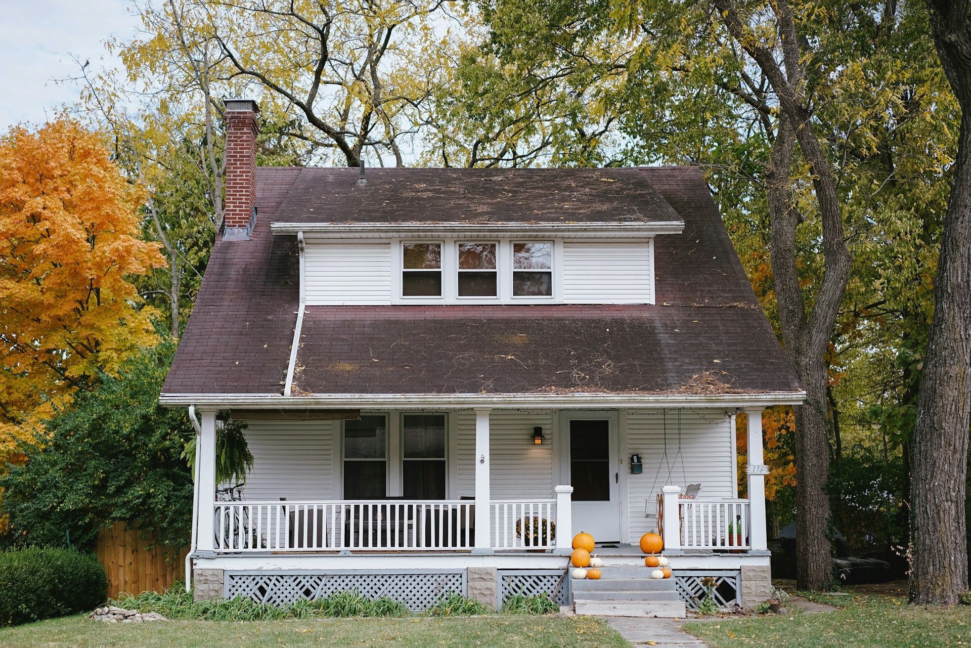 White two-story house with porch, chimney, and autumn trees; pumpkins on porch steps.