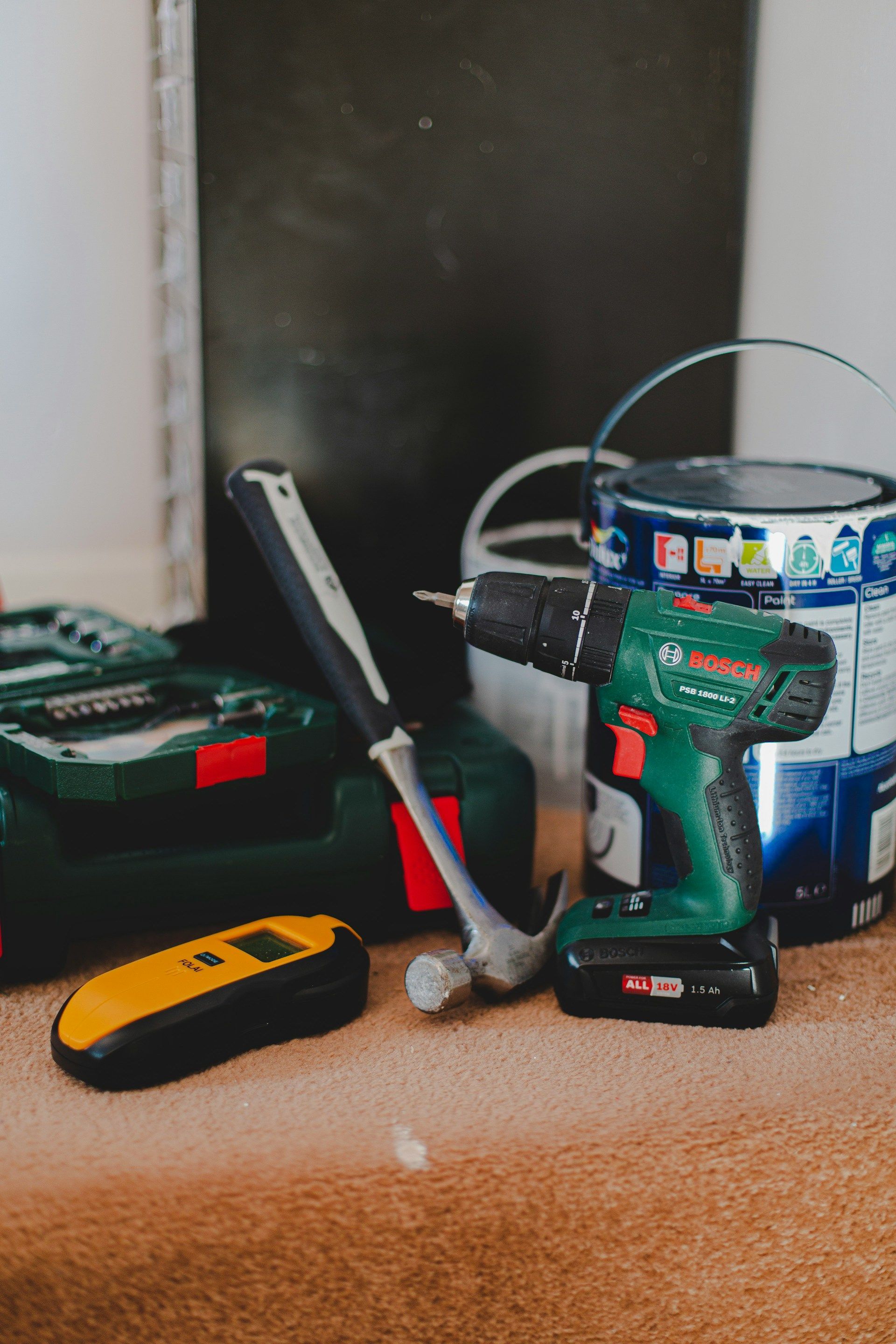 Tools on floor: drill, hammer, paint can, stud finder, and toolboxes; home repair setting.