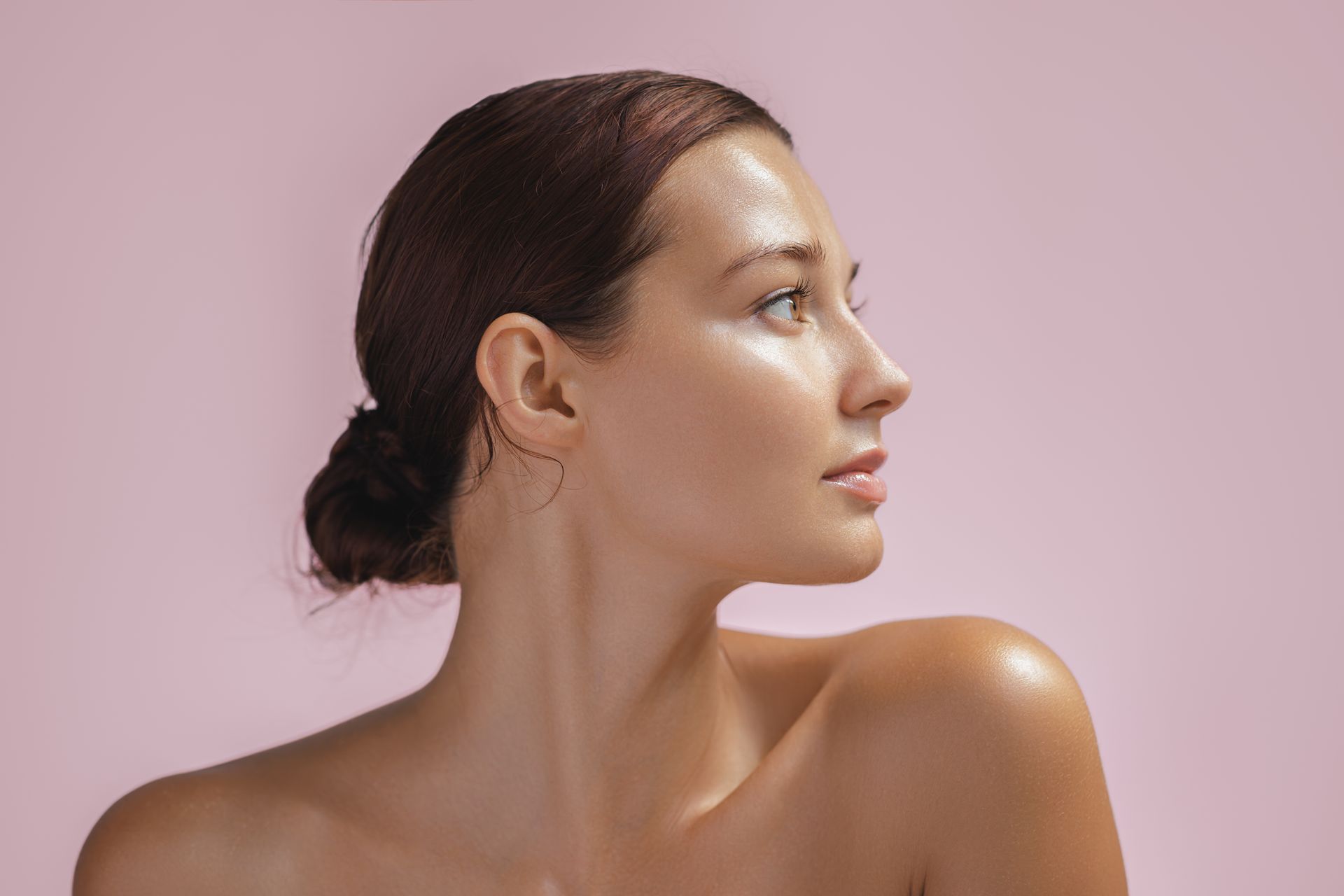 Woman with glowing skin, looking off-camera against a pink backdrop, hair pulled back in a bun.