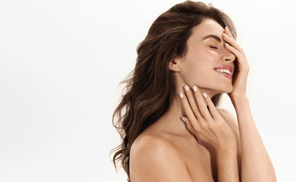 Woman with flowing dark hair smiles, shielding eyes with one hand, other hand near neck; white background.