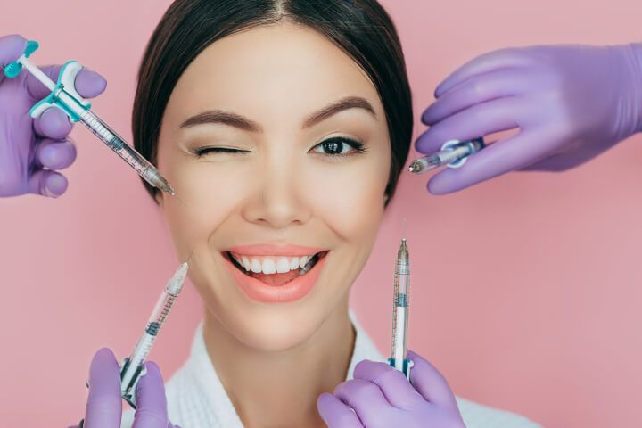 Woman winking, smiling, surrounded by hands holding syringes, on a pink background.