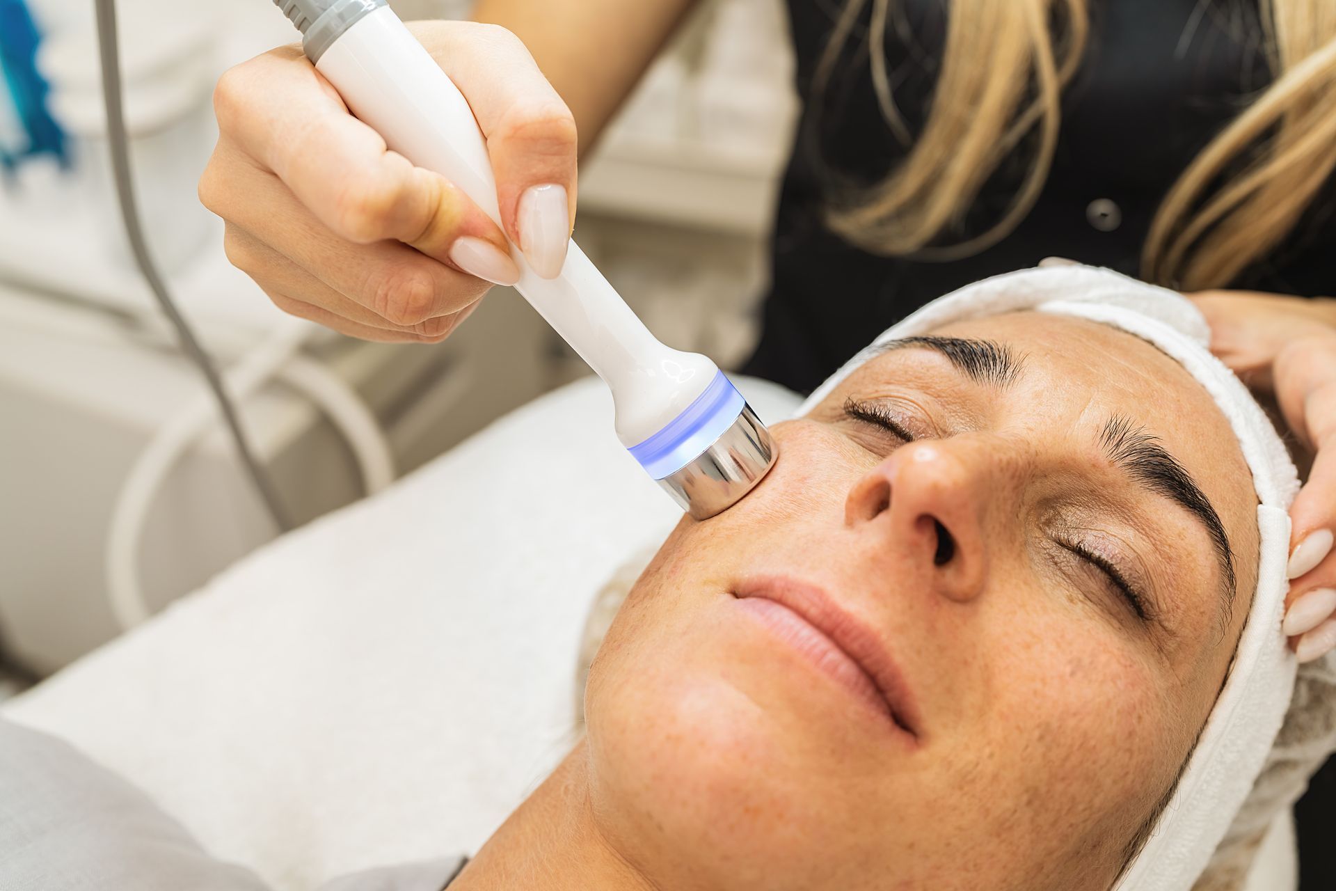 Woman receiving a facial treatment with a handheld device in a spa setting. The technician is holding the device to her face.