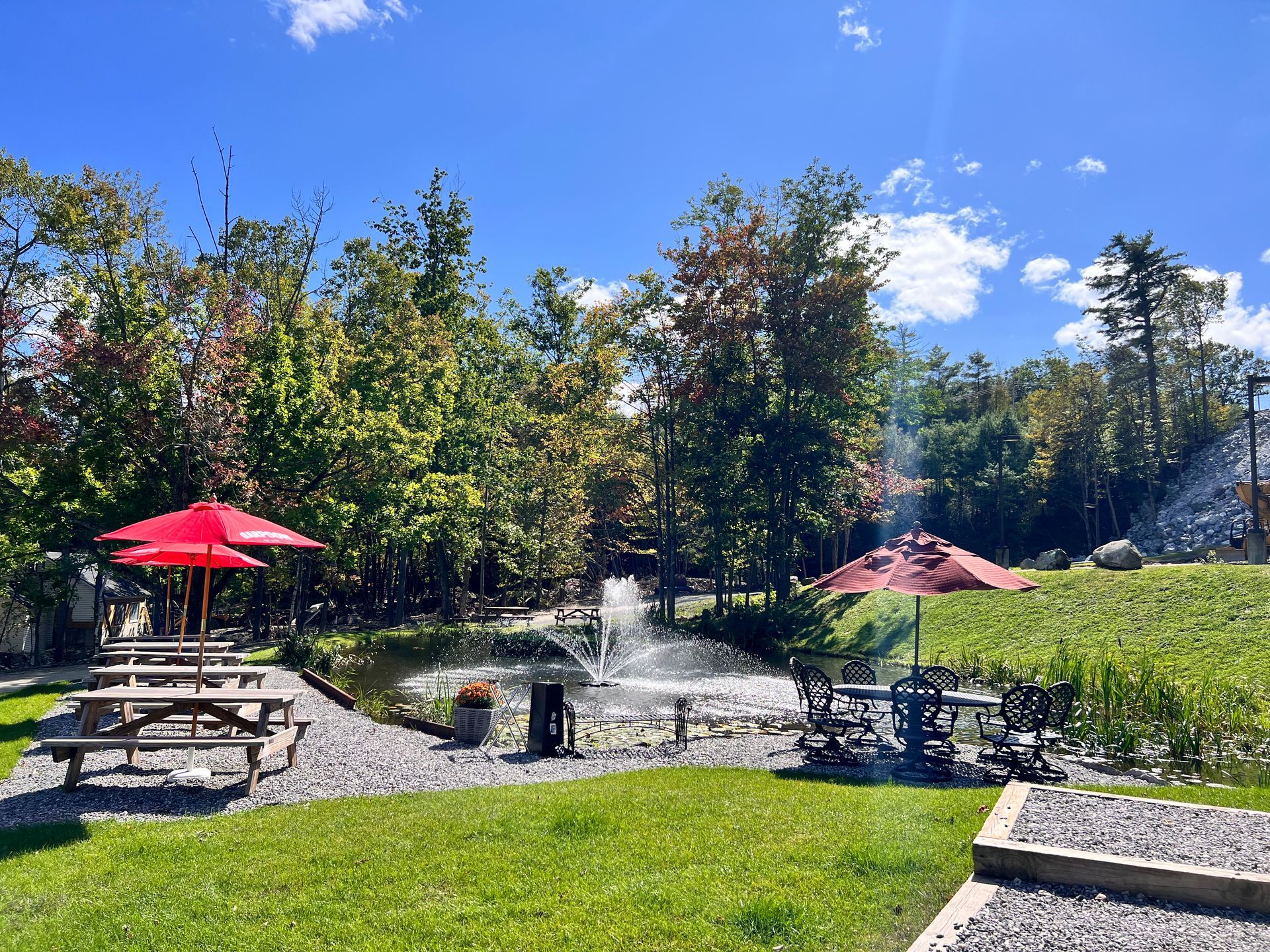 A park with a fountain , picnic tables , and red umbrellas.