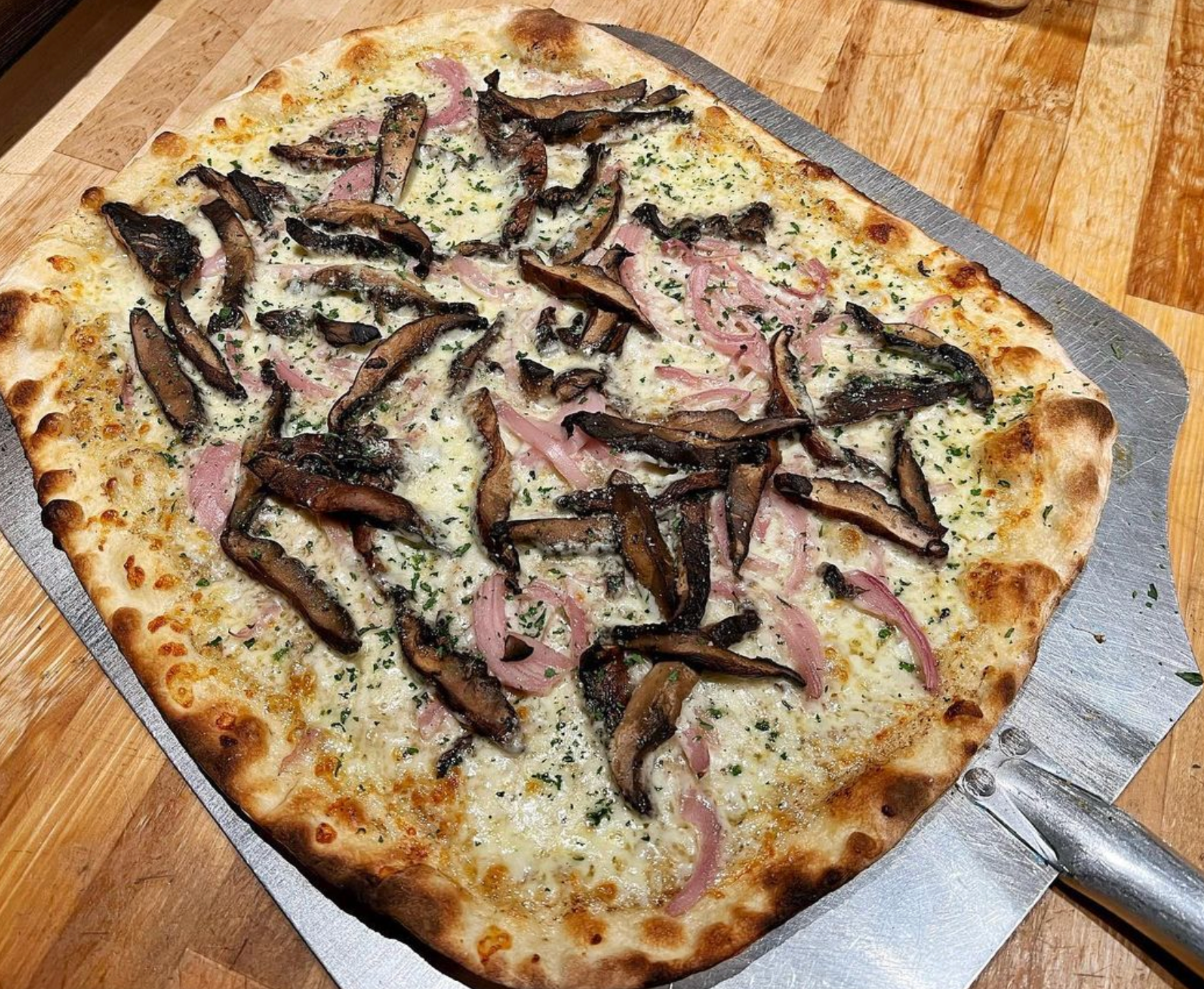 A pizza with mushrooms and ham on it is on a peel on a wooden table.
