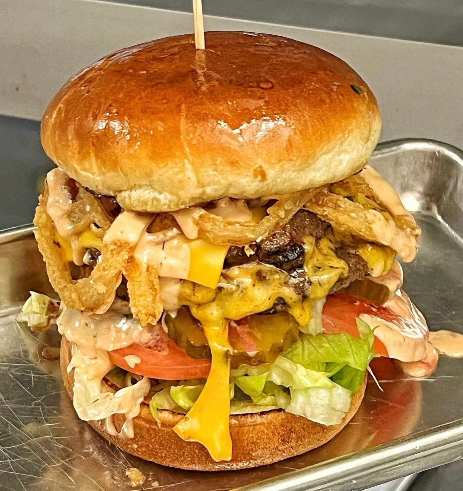 A hamburger with cheese , lettuce , onion rings and tomatoes on a bun on a metal tray.
