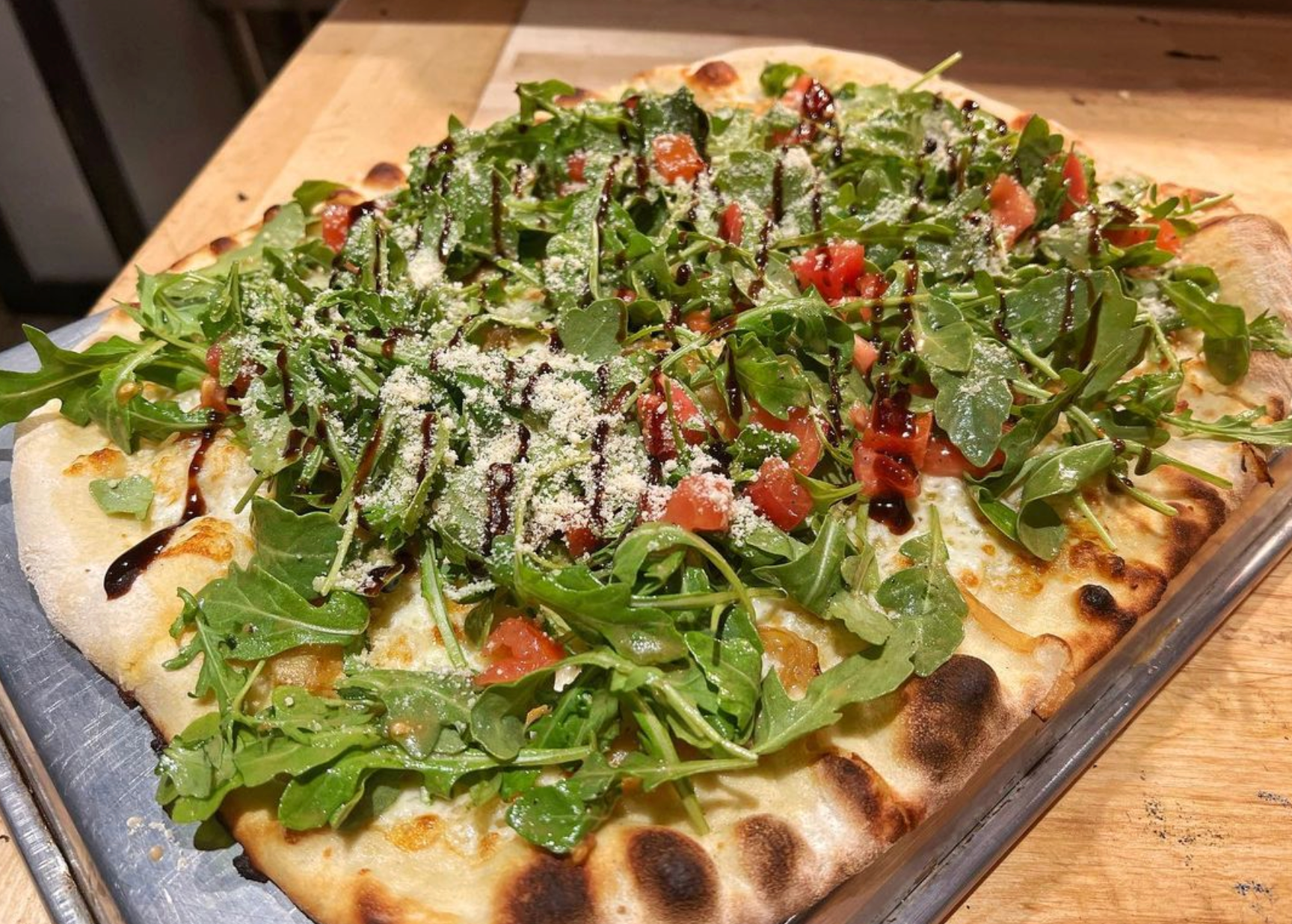 A pizza with tomatoes , greens , cheese and balsamic vinegar on a tray on a table.