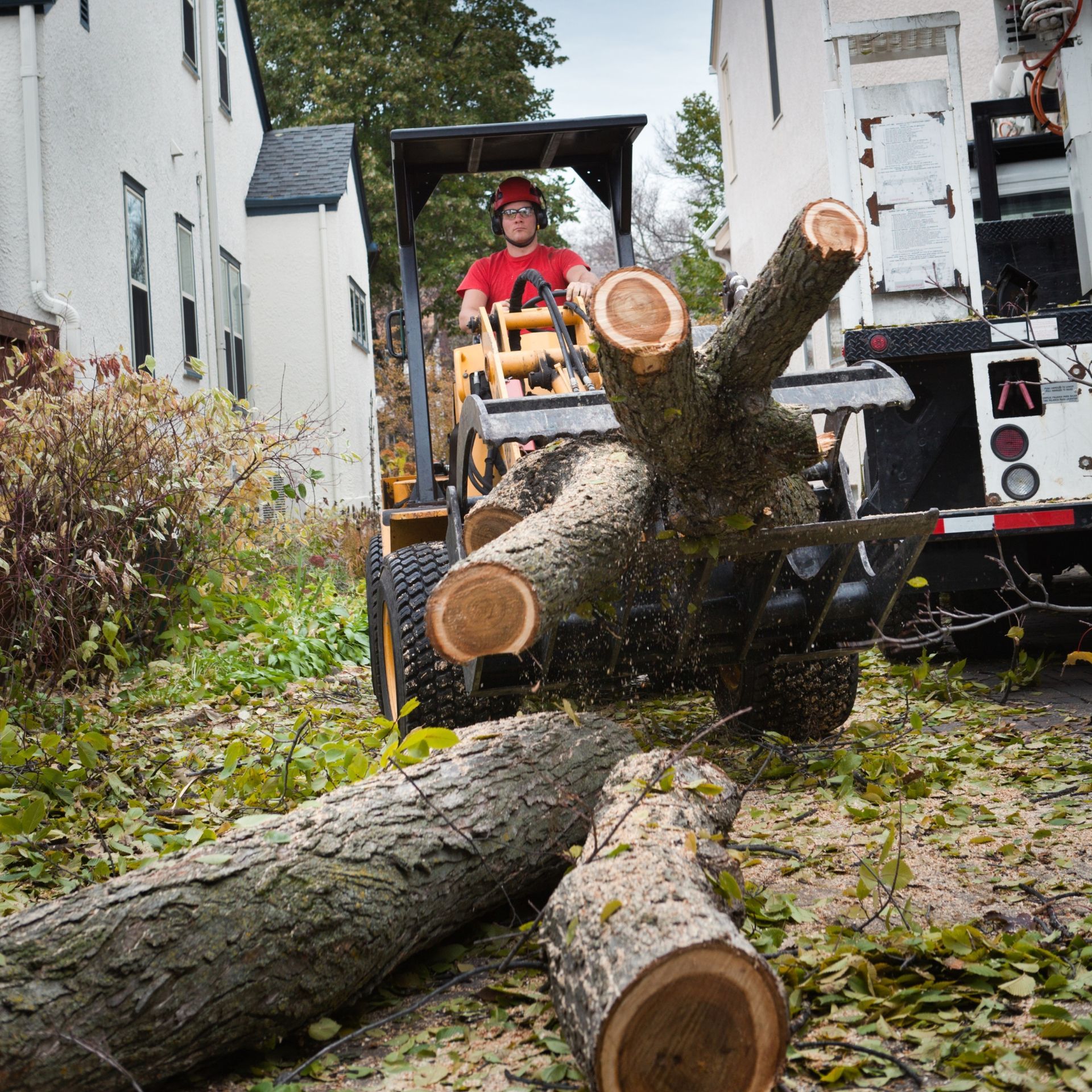 Man operating a yellow loader moving tree branches on a residential street.