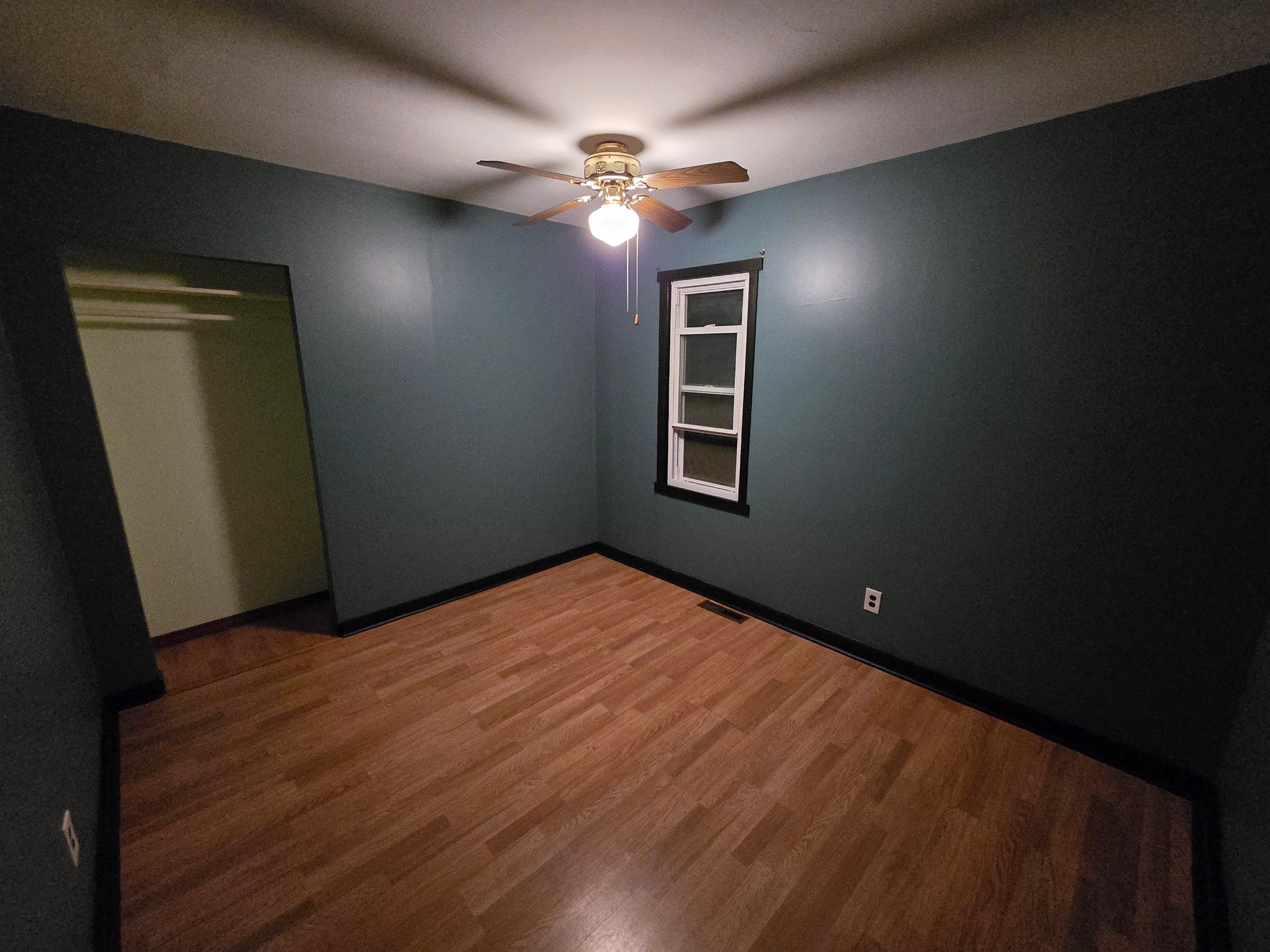 Empty bedroom with dark gray walls, wood floor, ceiling fan light, and a small window.