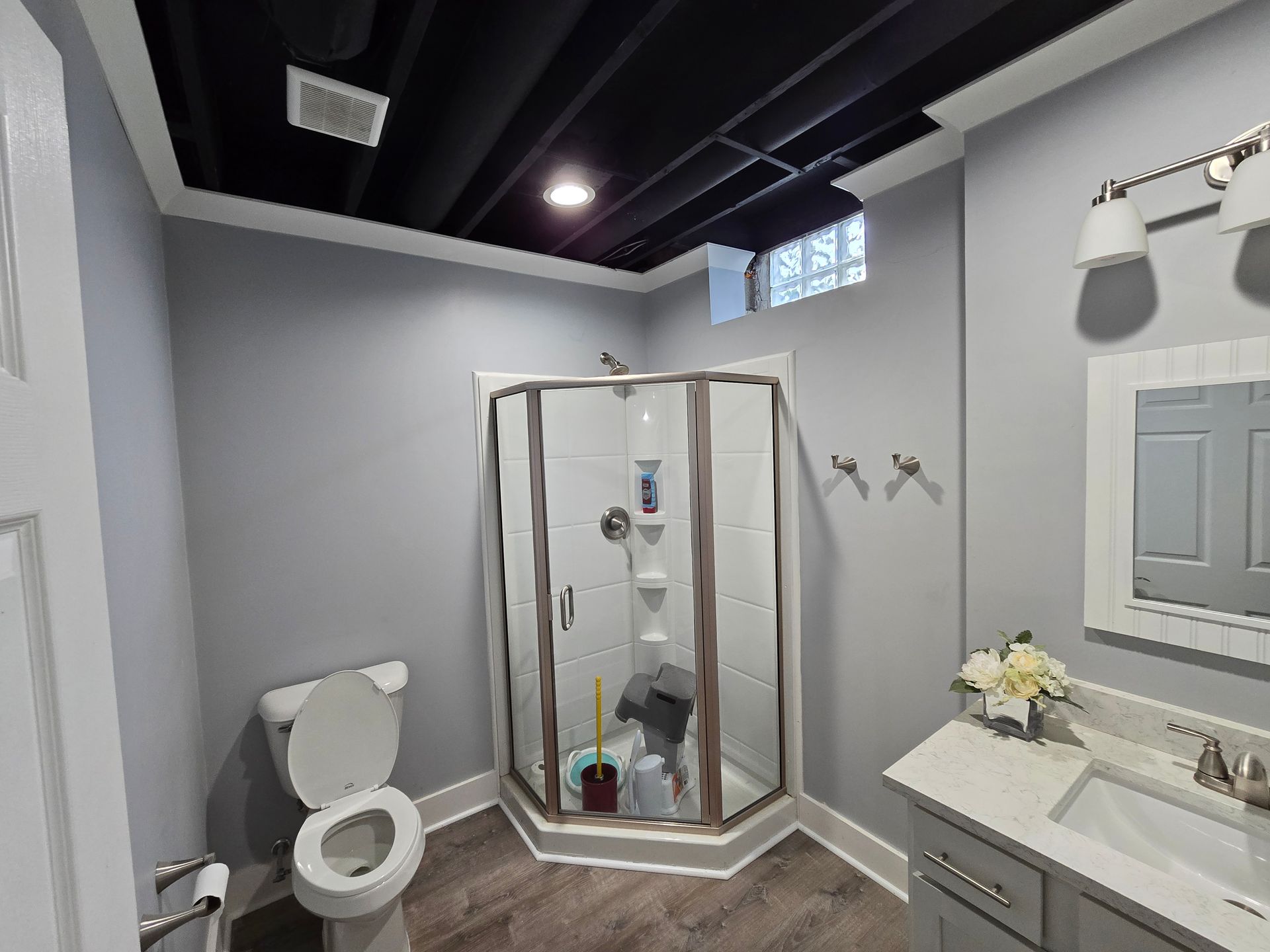 Small gray bathroom with shower stall, toilet, sink, mirror, and wall-mounted lights.