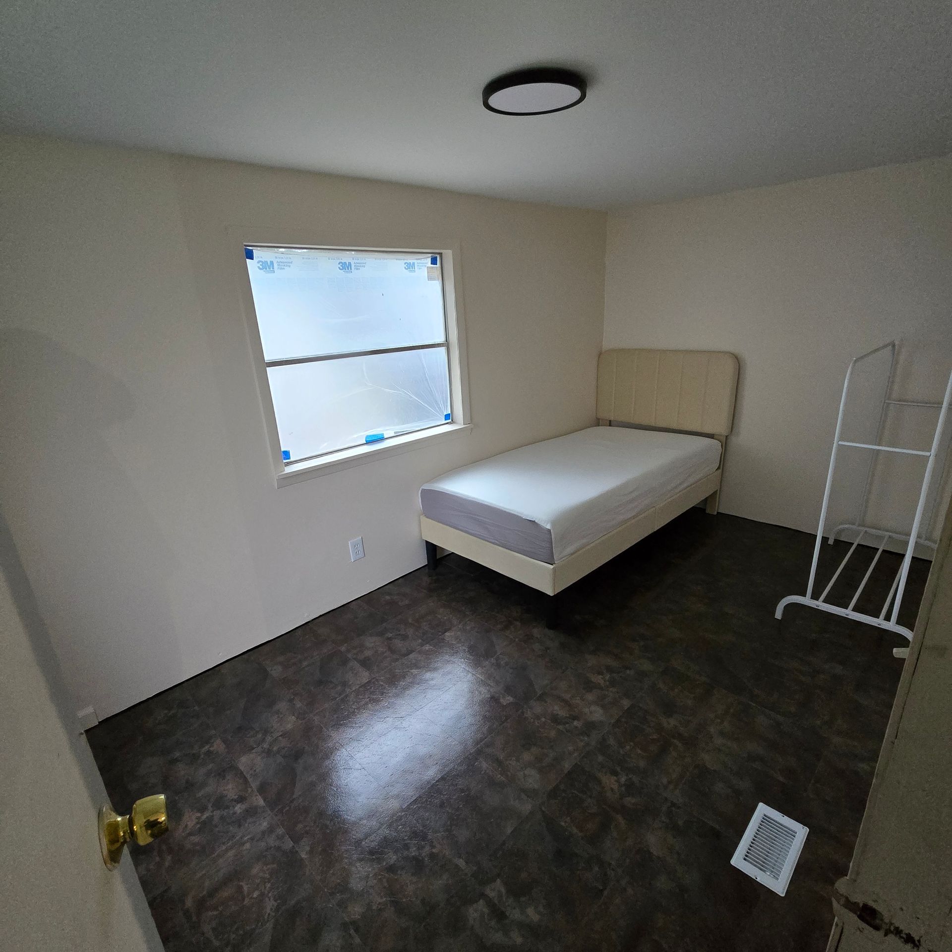 Small bedroom with a single bed, window, beige walls, dark floor, and a clothes rack.