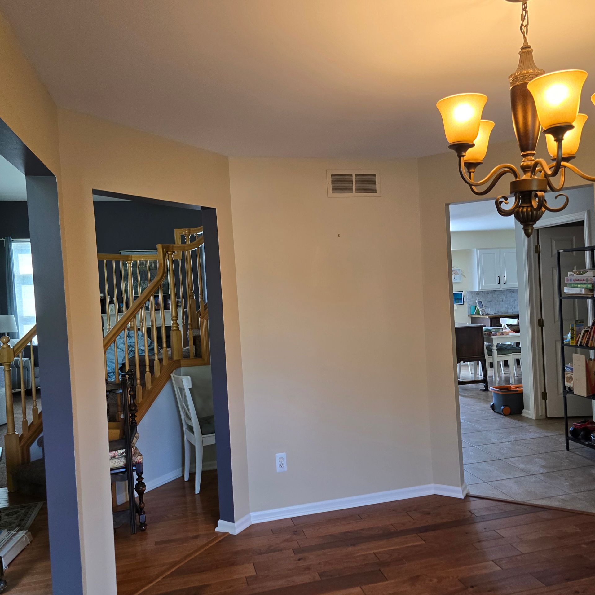 Empty dining room with hardwood floors, beige walls, staircase, and chandelier opening to a kitchen.