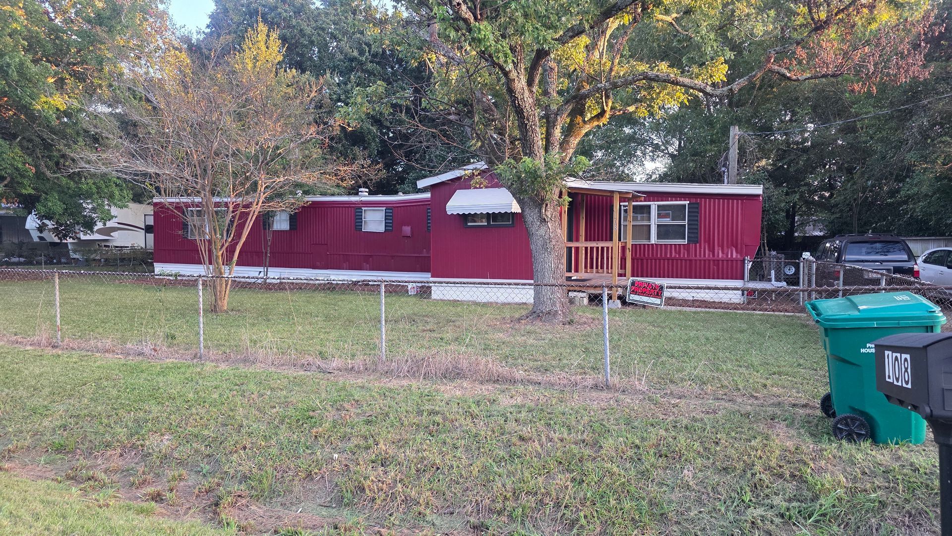 Red mobile home with white trim behind a grassy yard and chain-link fence, trees in the background.