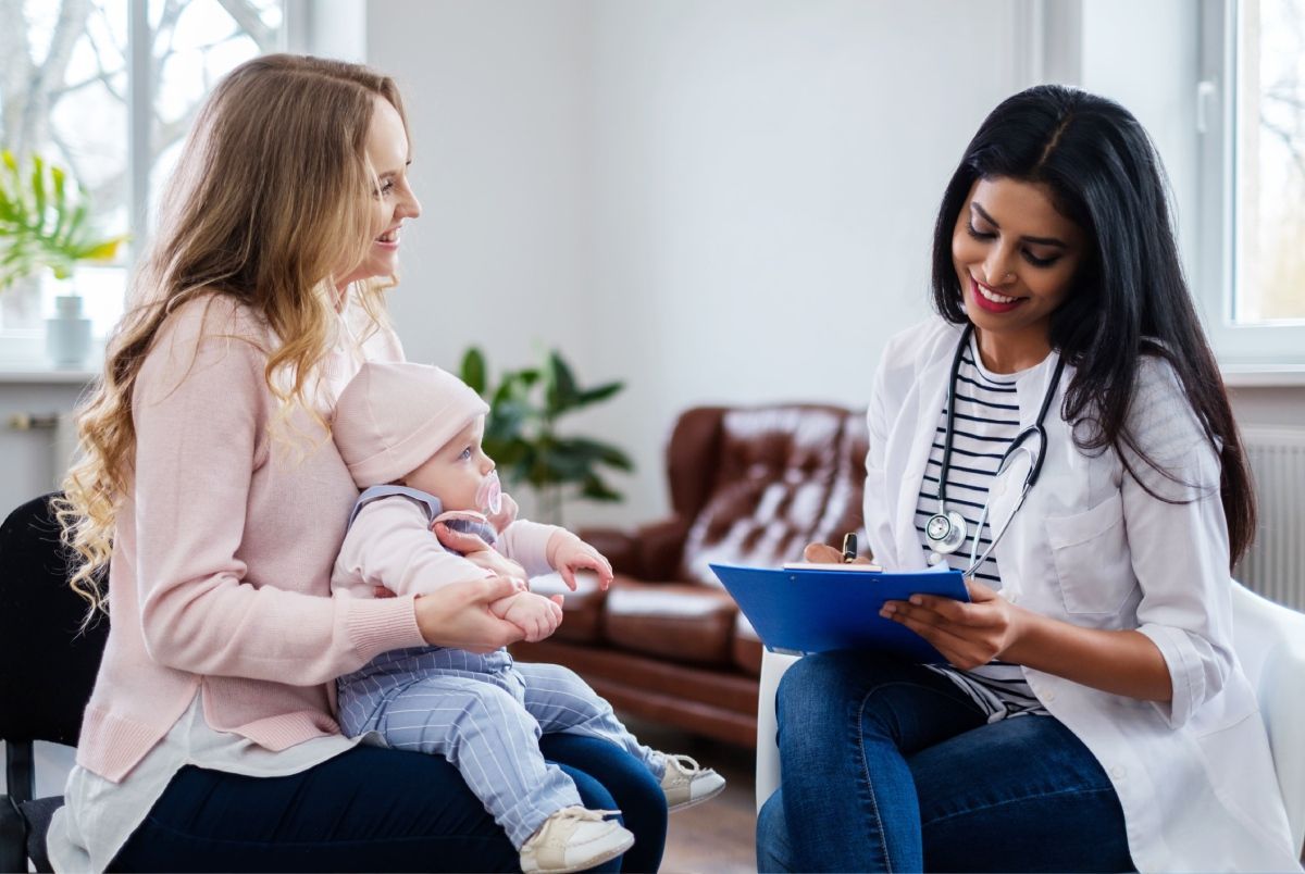 Mother and baby meet with a smiling clinician holding a blue clipboard in a bright living room