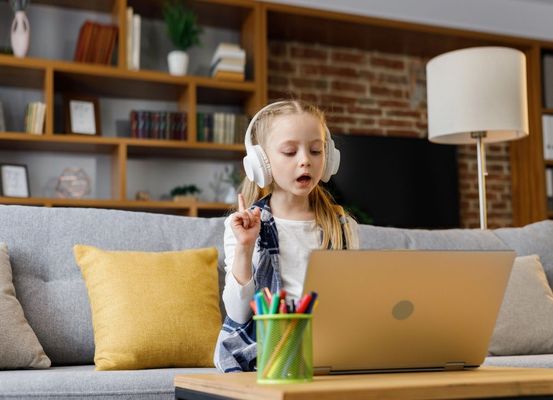 Child wearing headphones uses a laptop at home beside a pencil cup on a sofa.