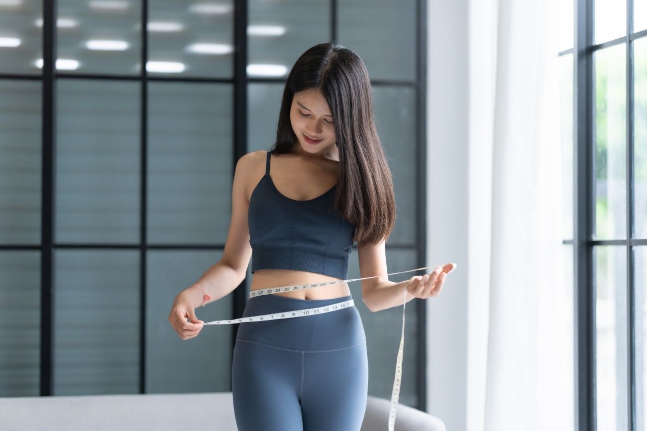 Woman standing indoors, smiling, in a black crop top and blue leggings by a bright window