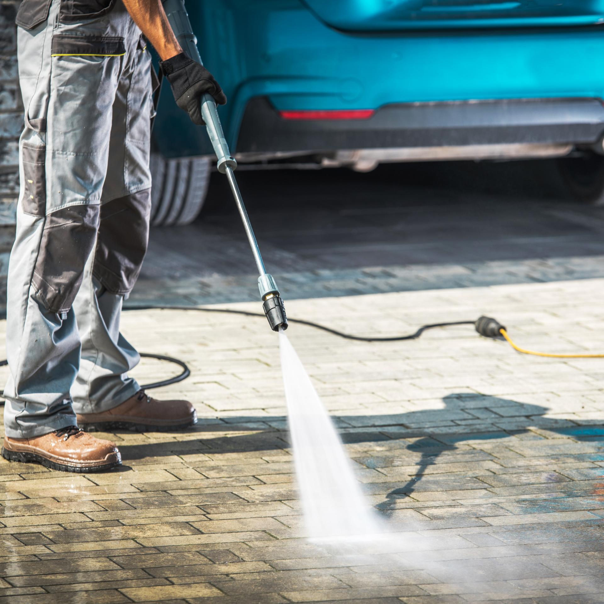 Person pressure washing a paved surface near a turquoise car.