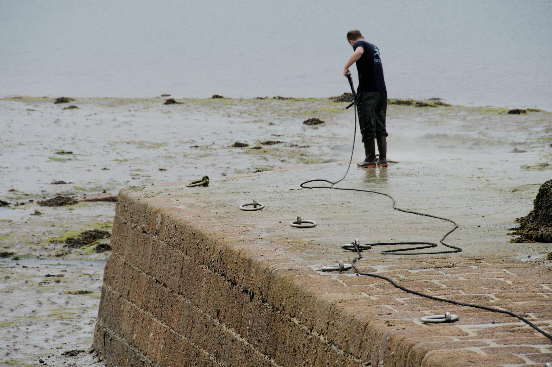 Man on a muddy shore, wearing boots, holding a tool, with a long cable connected to anchors on a stone wall.