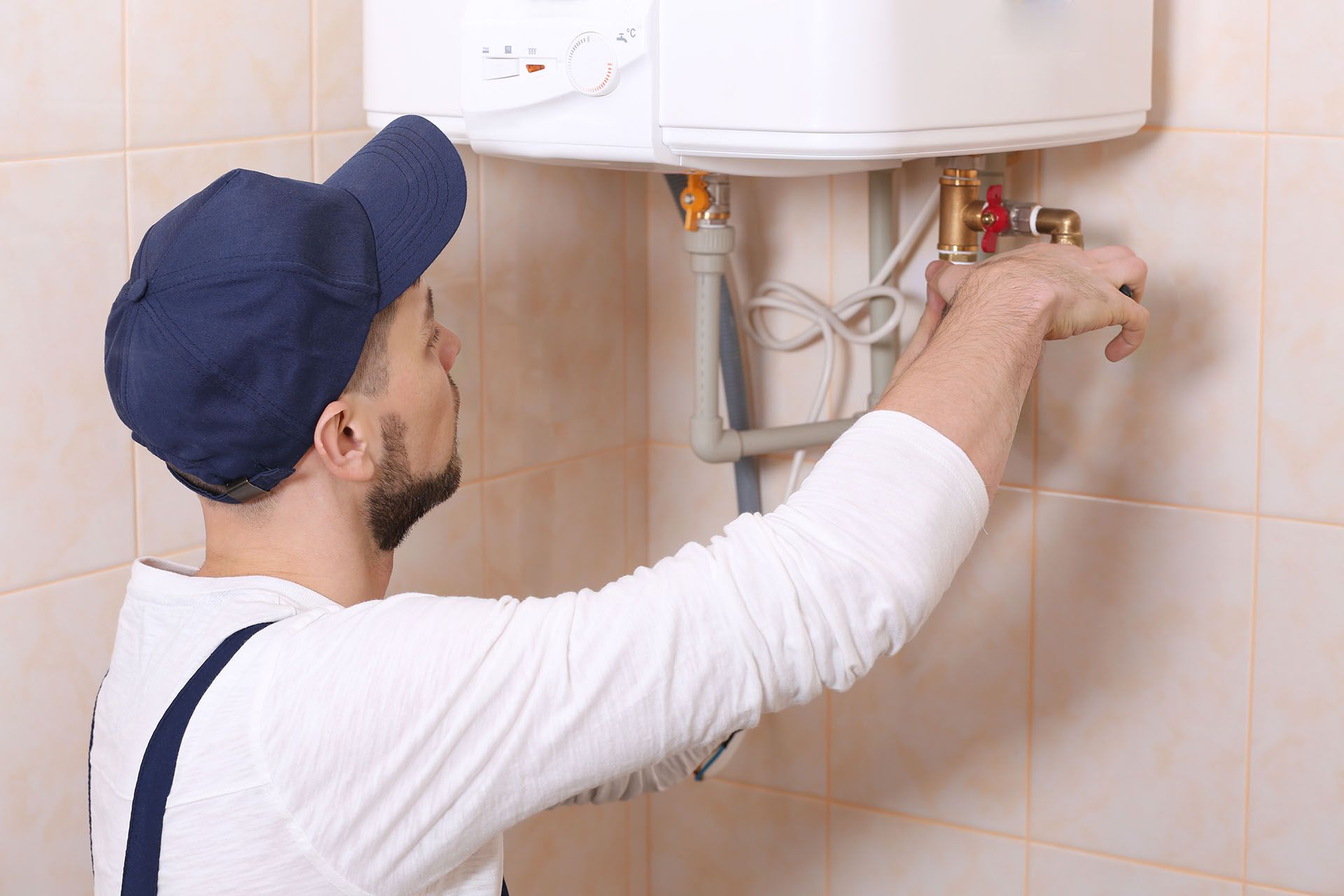 A plumber in a blue cap working on a white water heater in a tiled room