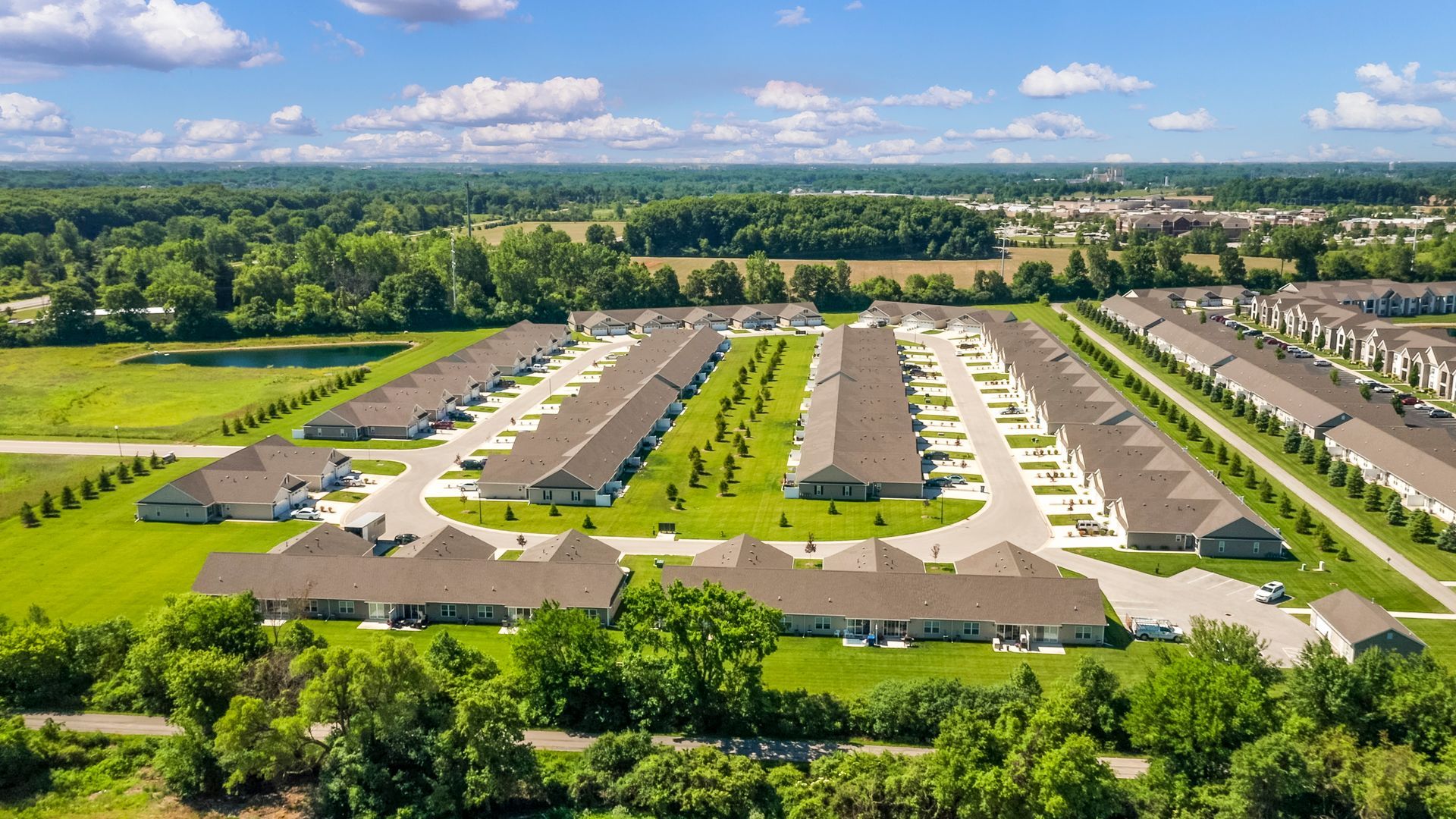Aerial view of a suburban neighborhood with rows of houses, green lawns, and trees, under a blue sky.