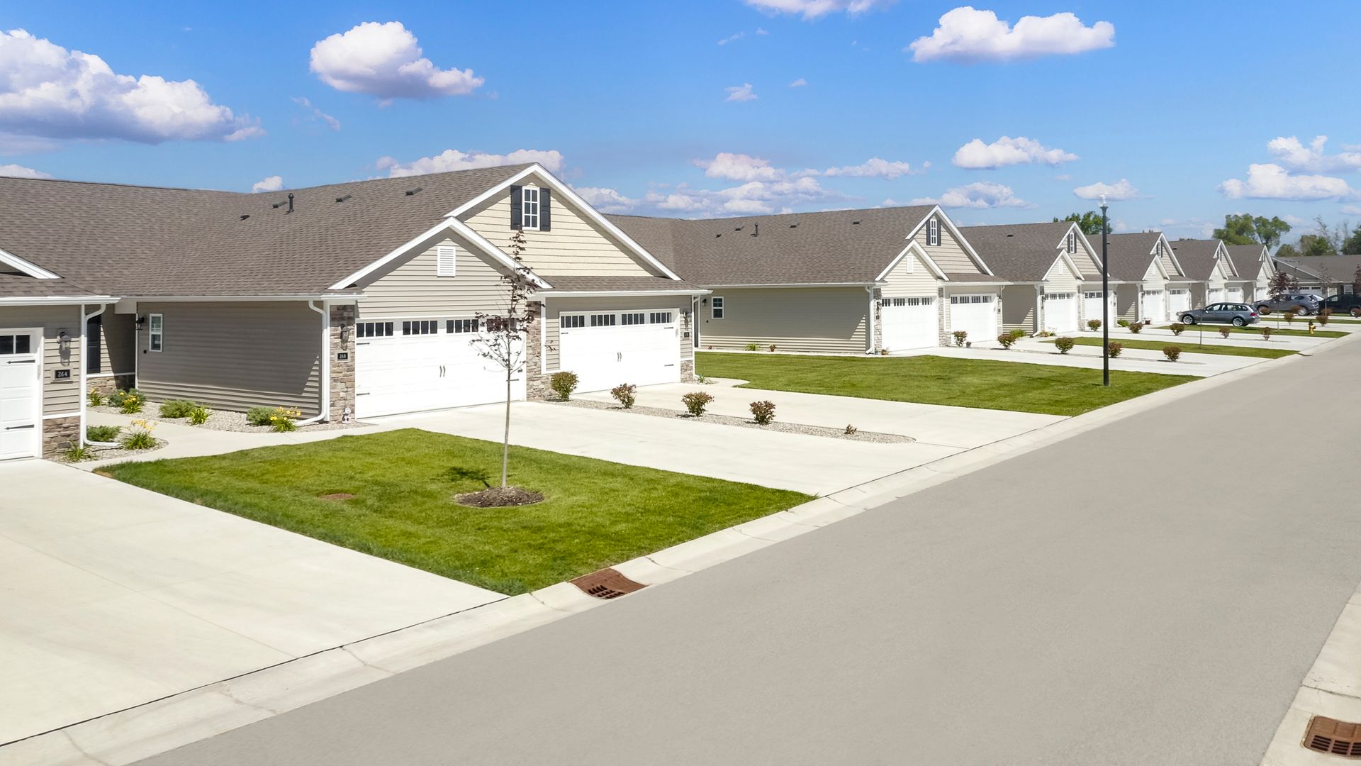 Row of beige houses with white garages and green lawns along a paved street under a blue sky.