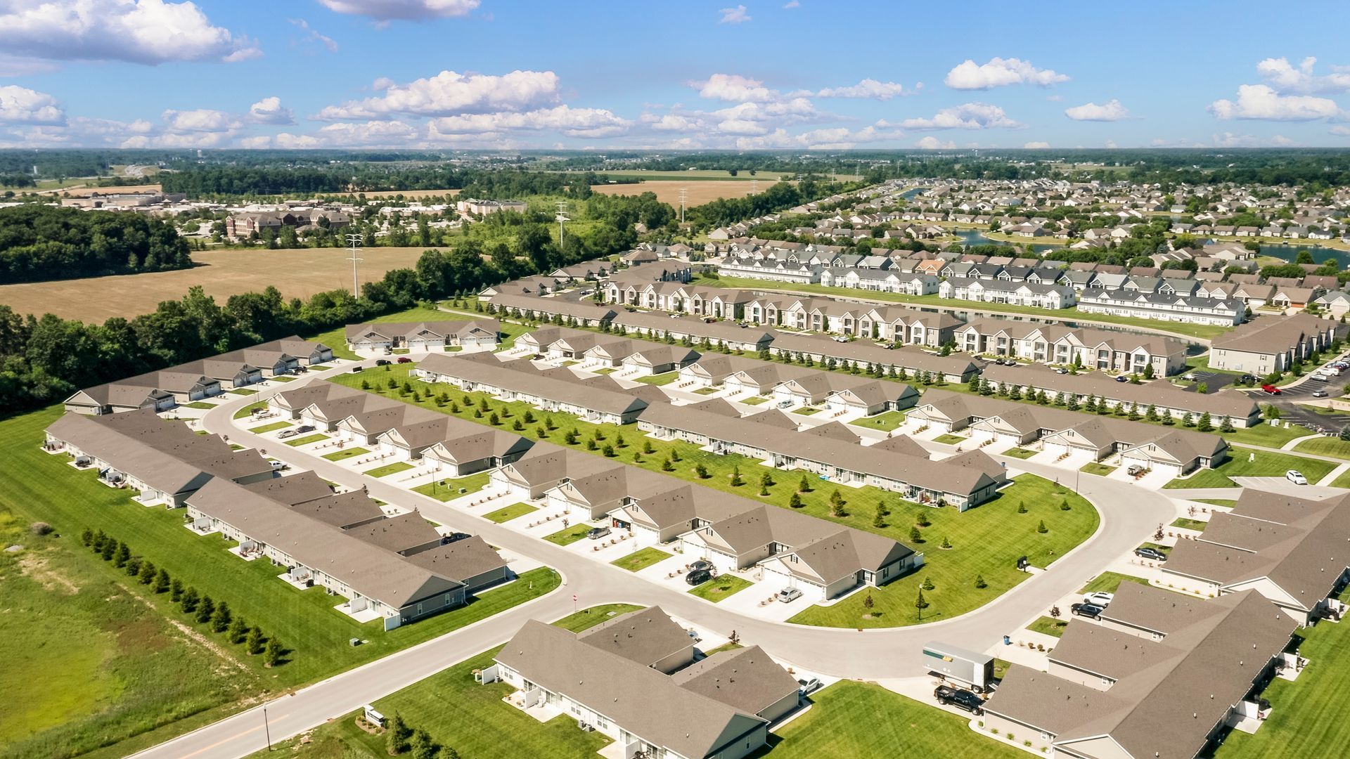 Aerial view of a suburban neighborhood with rows of houses, green lawns, and trees, on a sunny day.