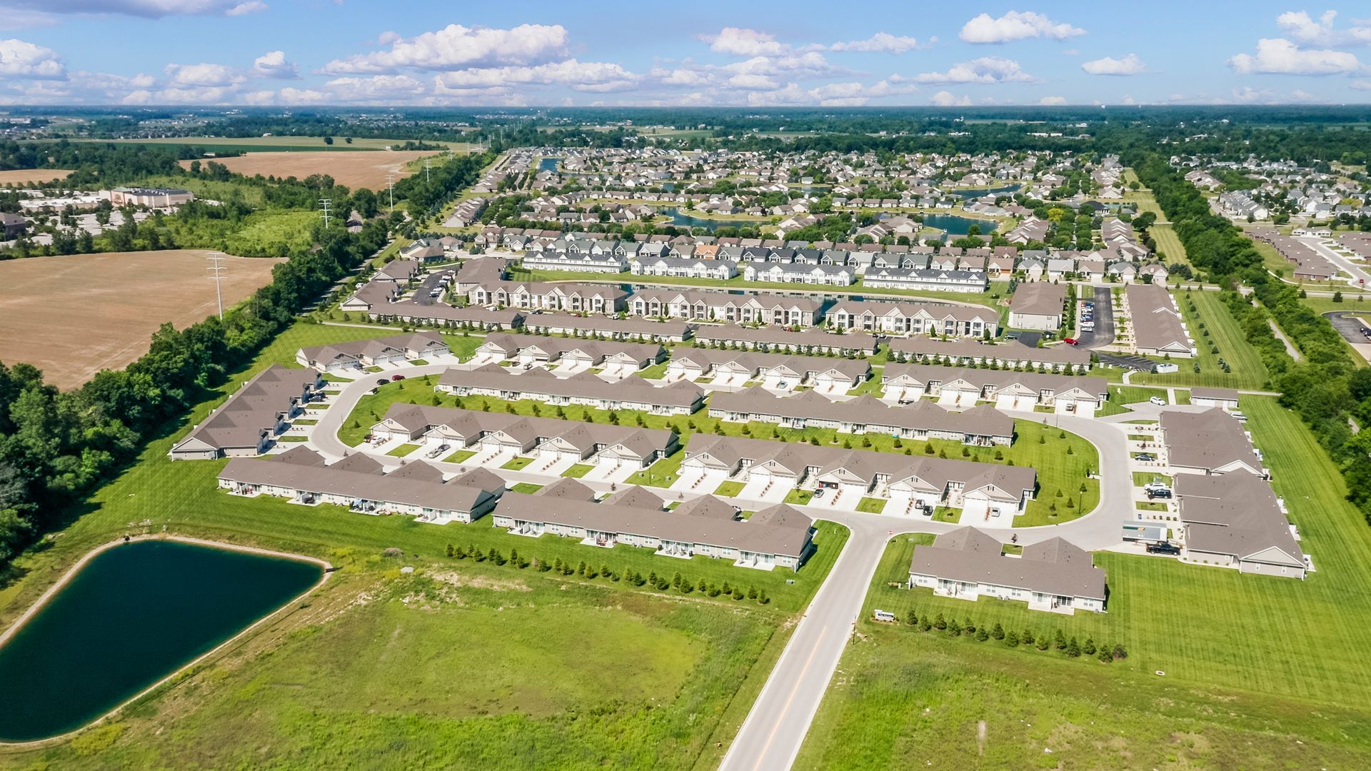 Aerial view of a suburban neighborhood with rows of houses, a lake, and green fields under a blue sky.