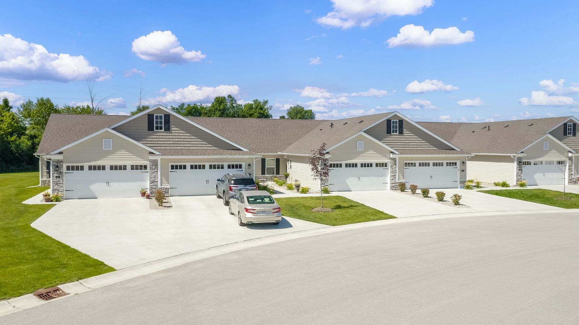 Row of light gray townhomes with attached garages on a sunny day. Paved driveways and lawn in front.