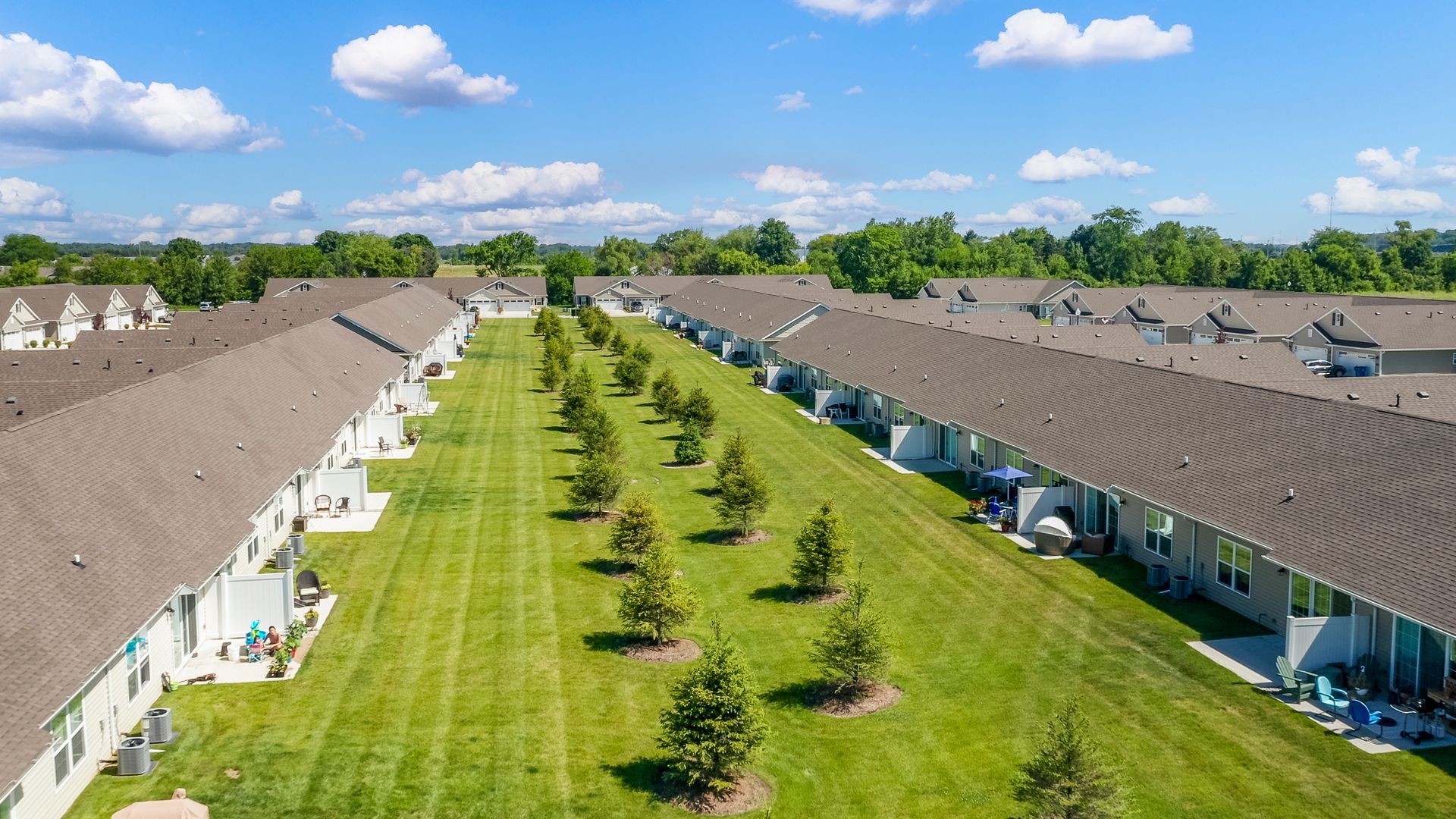 Aerial view of a row of townhomes with brown roofs, green lawns, and small trees on a sunny day.