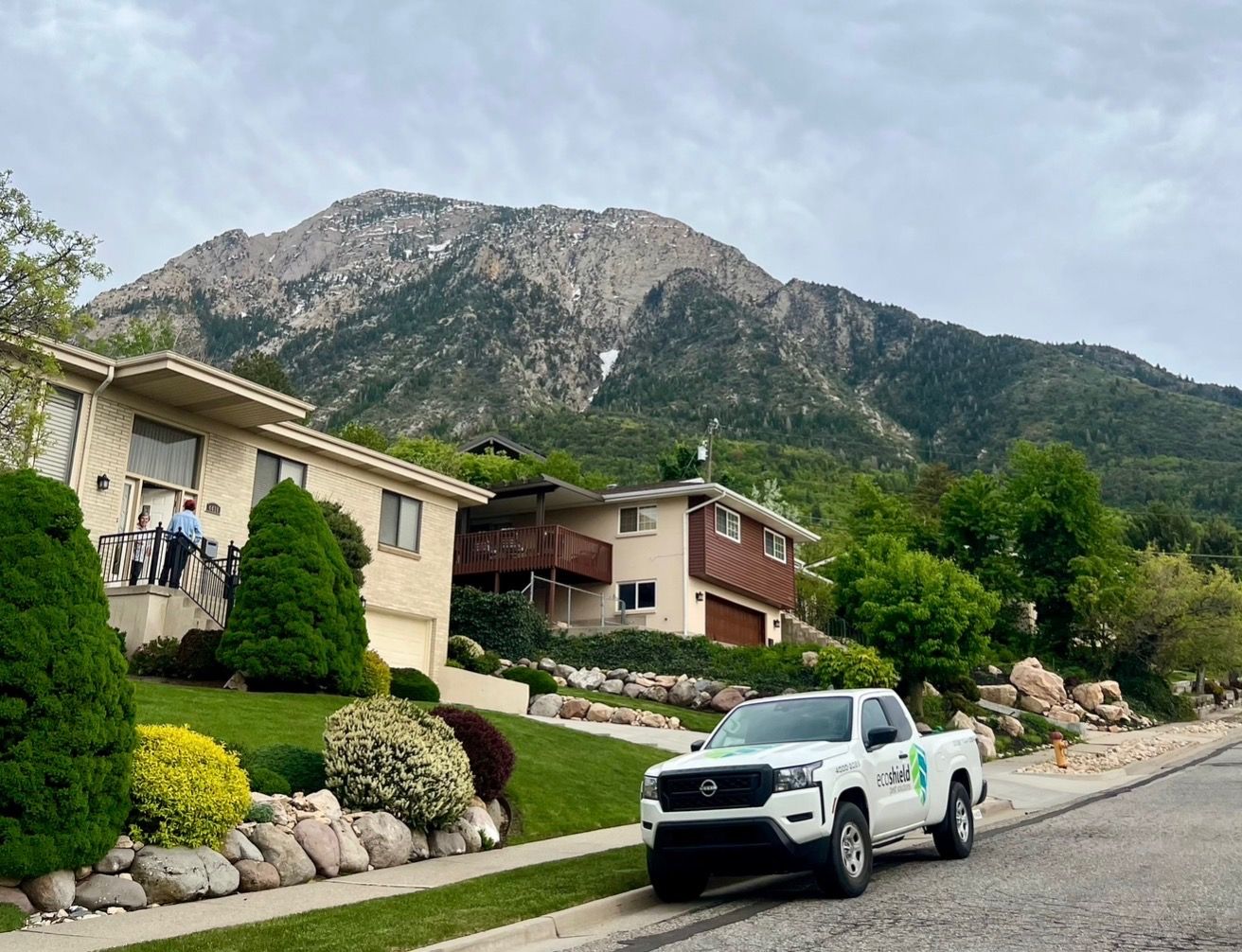 A smiling man drives an EcoShield pest solutions truck in a residential neighborhood on a sunny day.