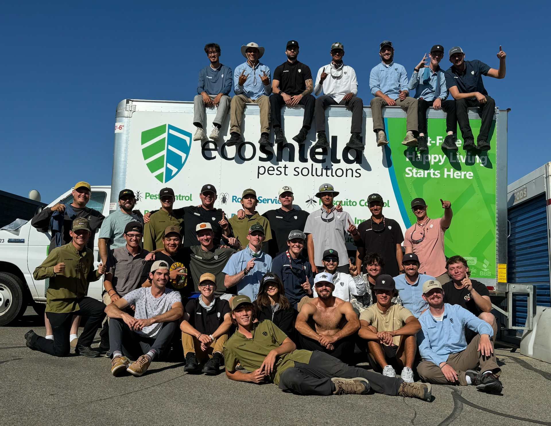 Pest control worker standing by truck, holding sprayer. He wears a cap, khaki pants, and a backpack, near a logo.