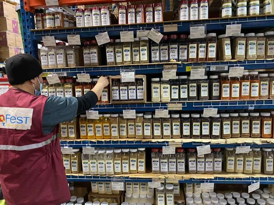 A man in a red vest is standing in front of a shelf filled with jars of spices.