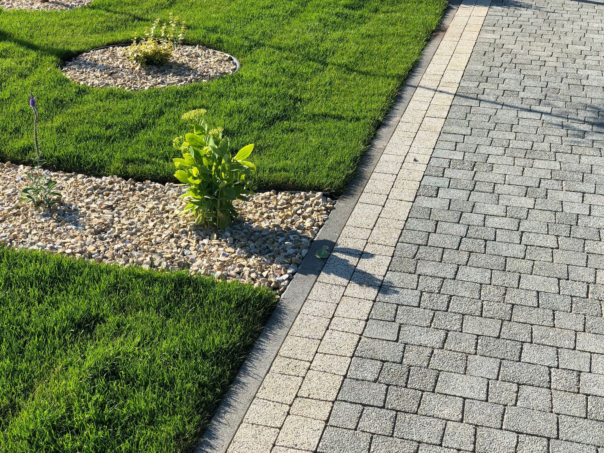 A landscaped area featuring a green lawn, a gravel border with small plants, and a gray cobblestone path.