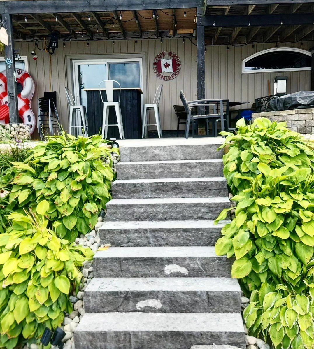 Stone steps lead to a patio with white chairs, bar, and Canadian flag. Lush green plants flank the steps.
