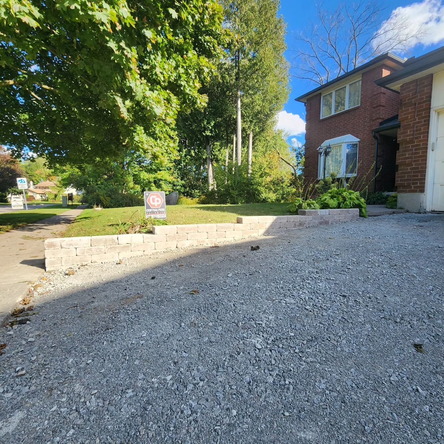 Gravel driveway leading to a brick house with a low stone wall and lawn. Trees and blue sky in background.
