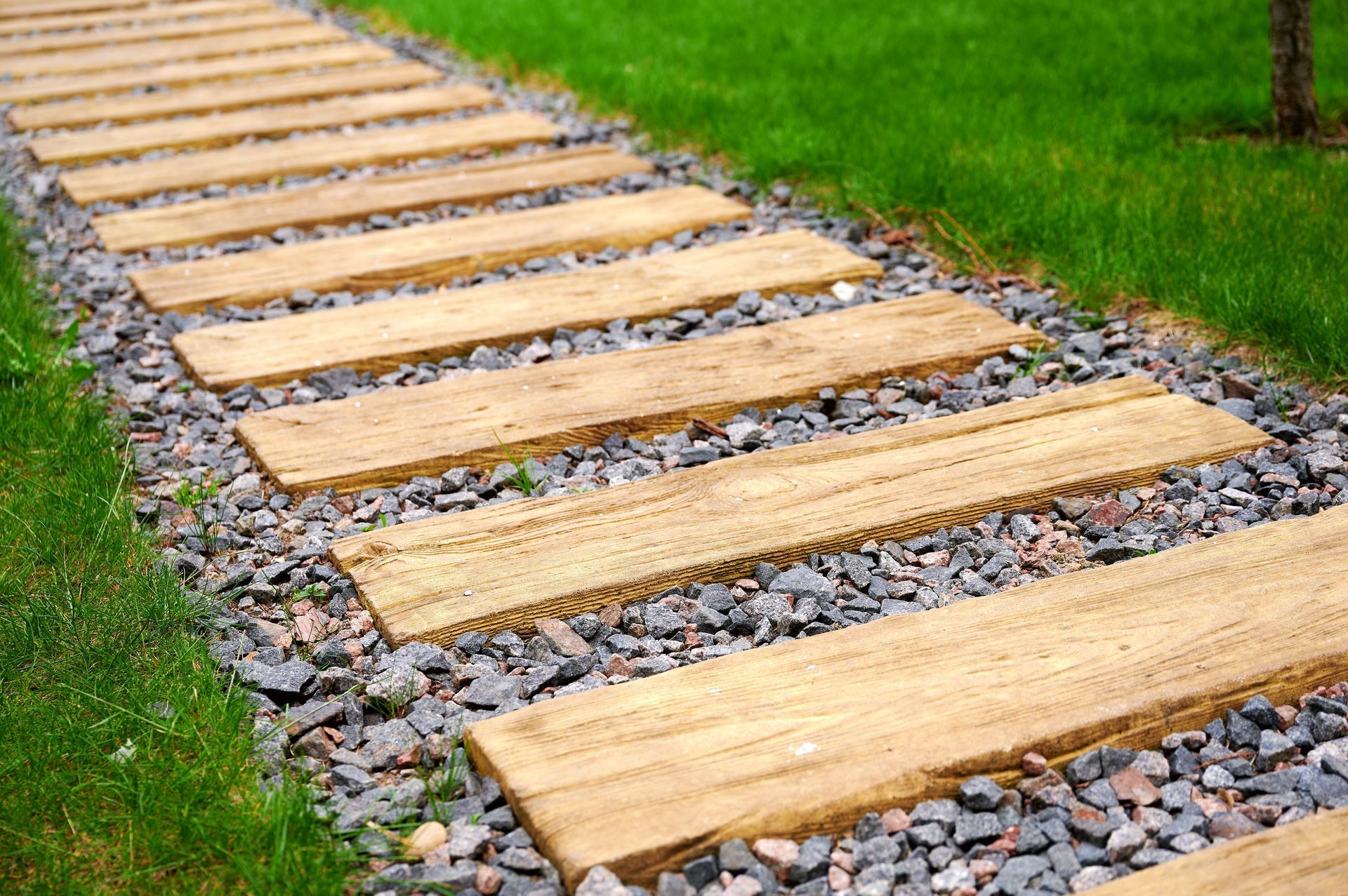 A gravel garden path features a series of horizontal, wood-grain stepping stones set between strips of green grass.