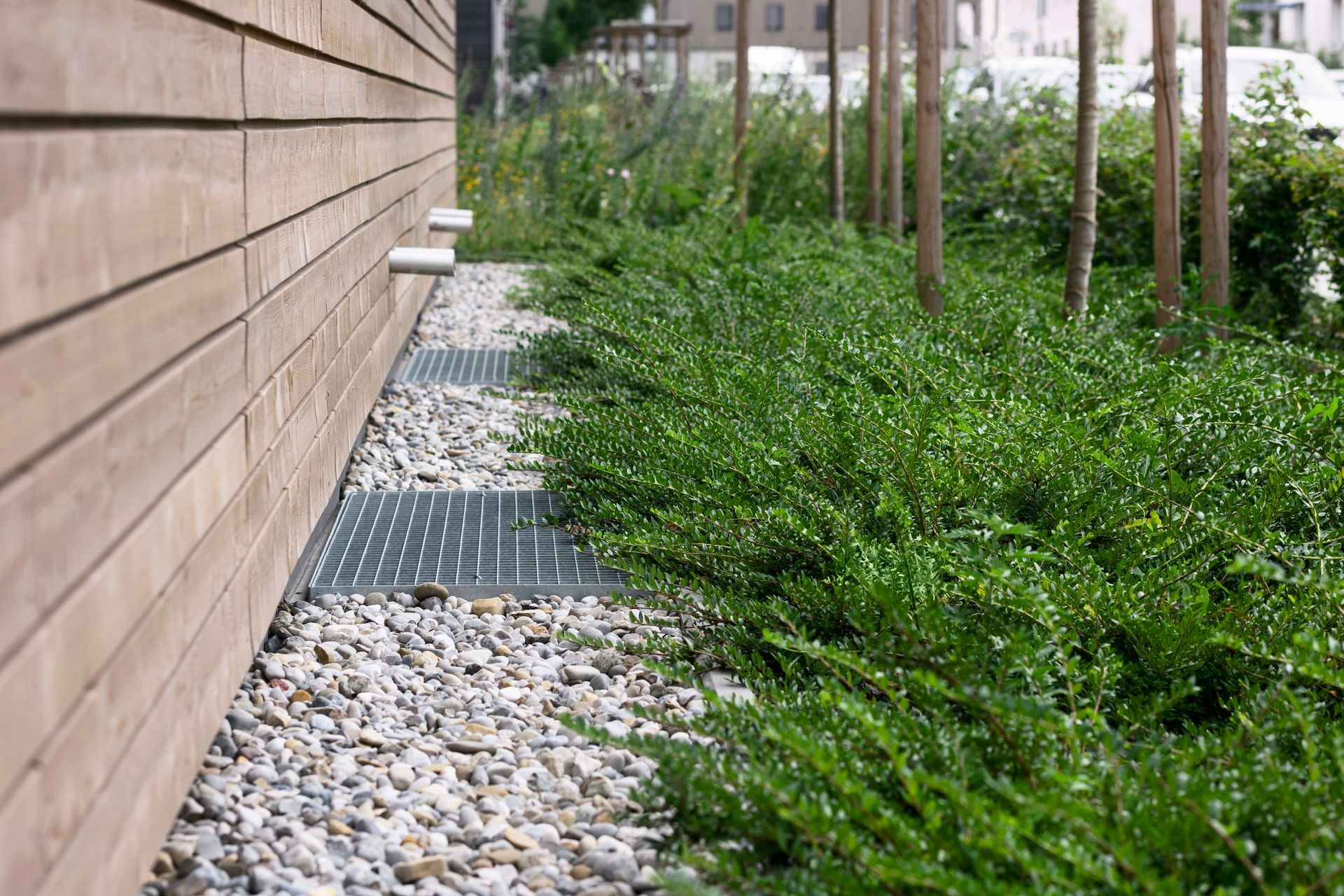 A gravel drainage strip with metal grates along a wooden-sided building, bordered by a dense green hedge.