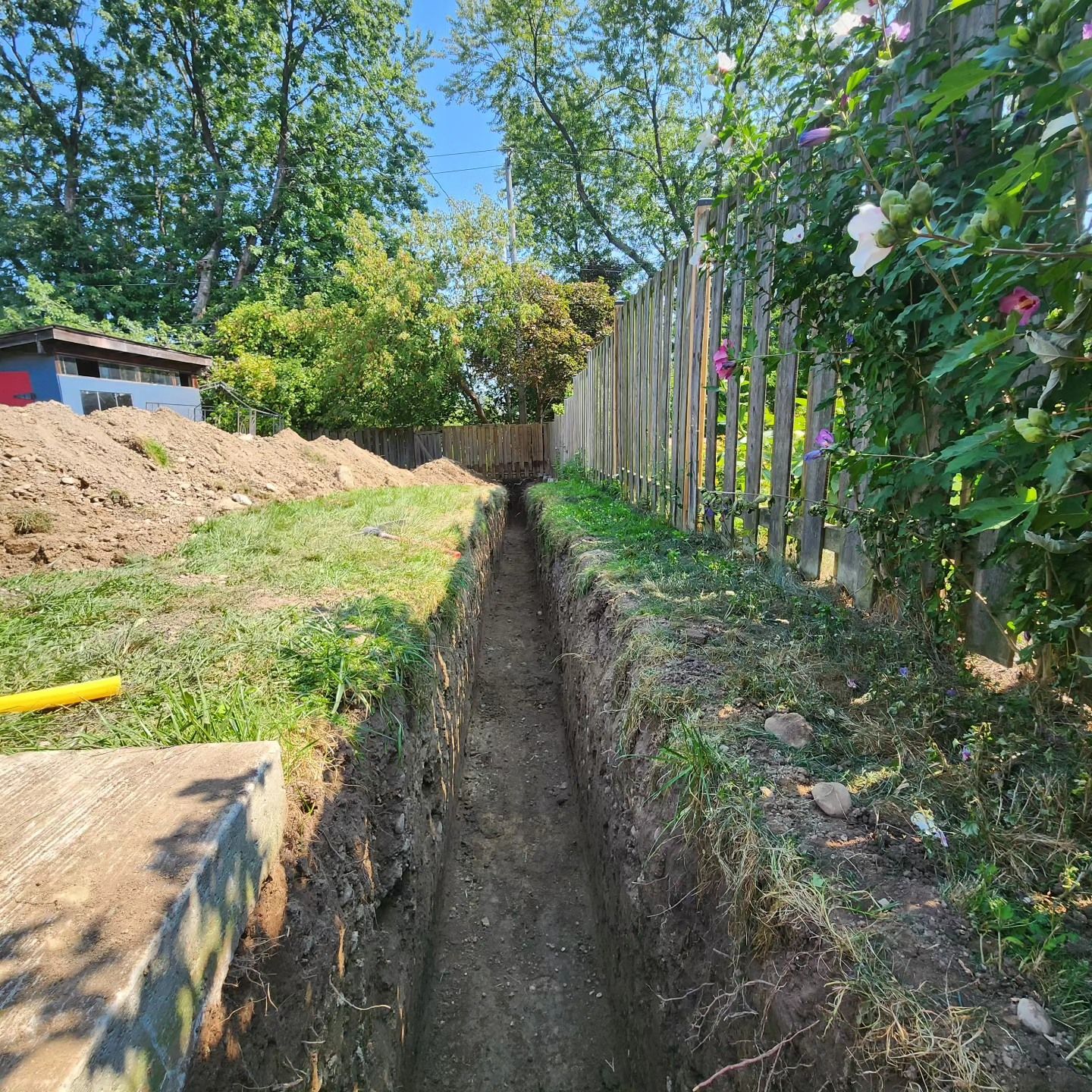 A trench is being dug in the grass next to a wooden fence.