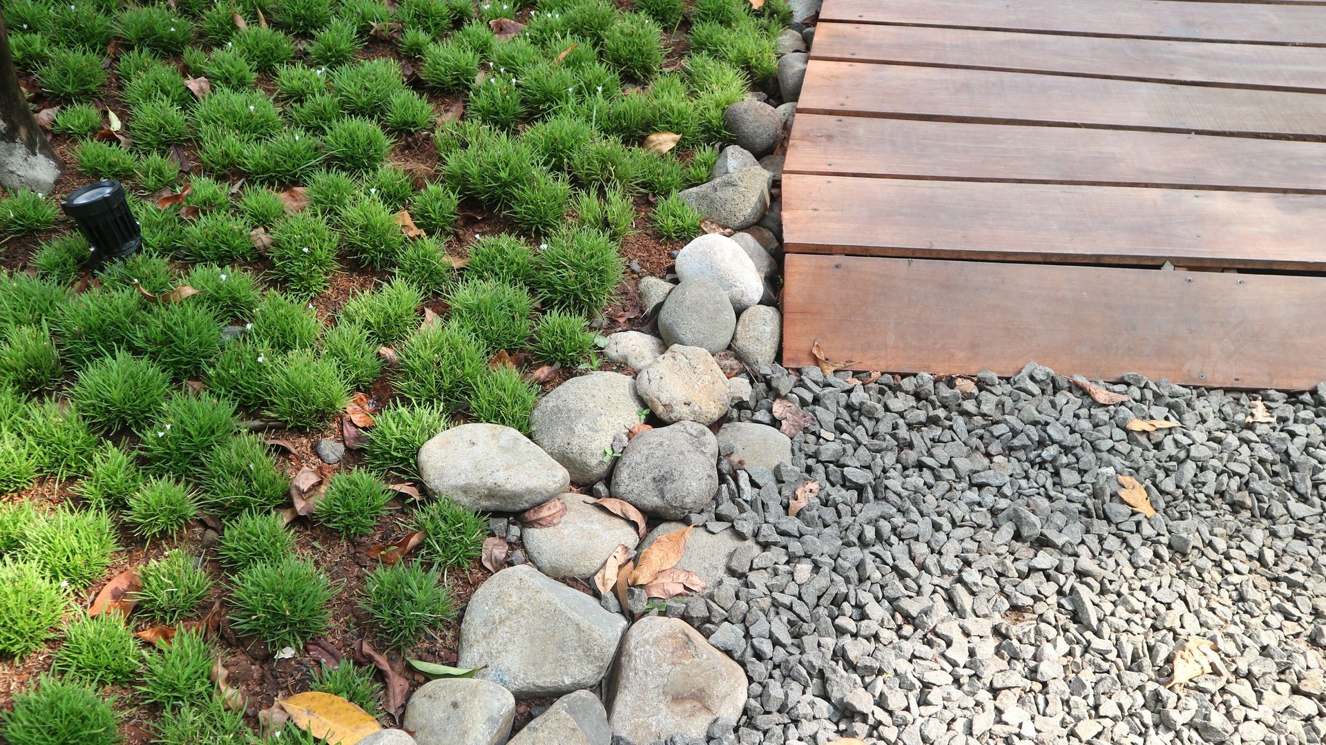 A wooden walkway borders a gravel path, separated by a line of smooth stones alongside a bed of small, green plants.