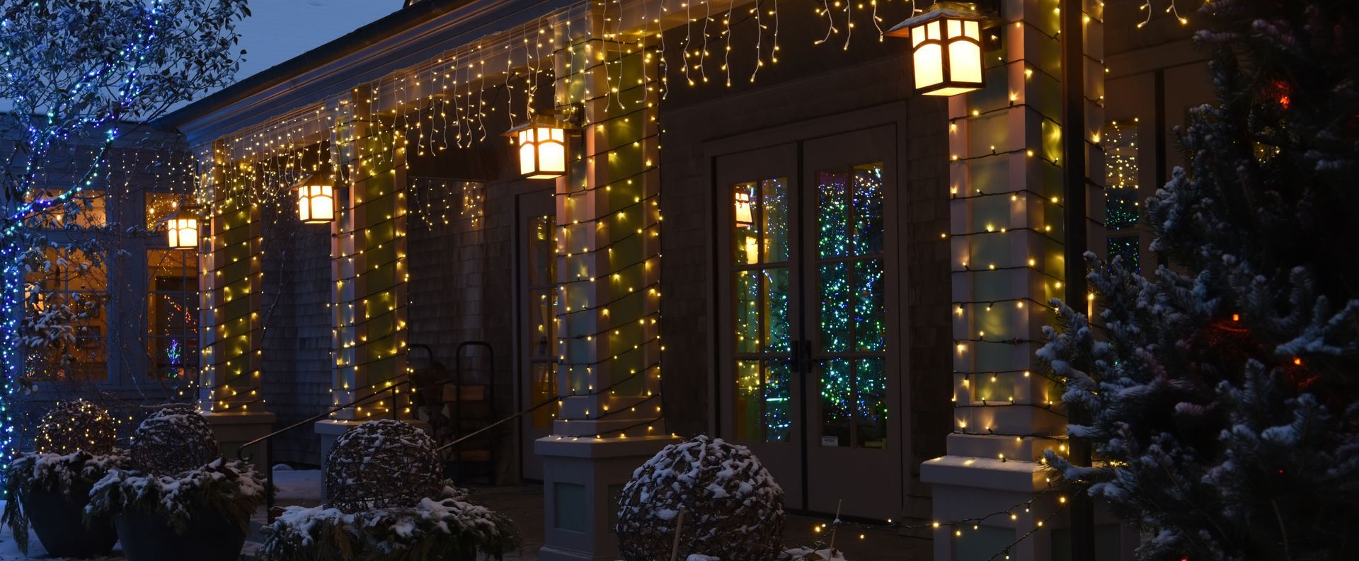 A house entrance decorated with yellow and blue Christmas lights and lanterns, snow on the bushes.