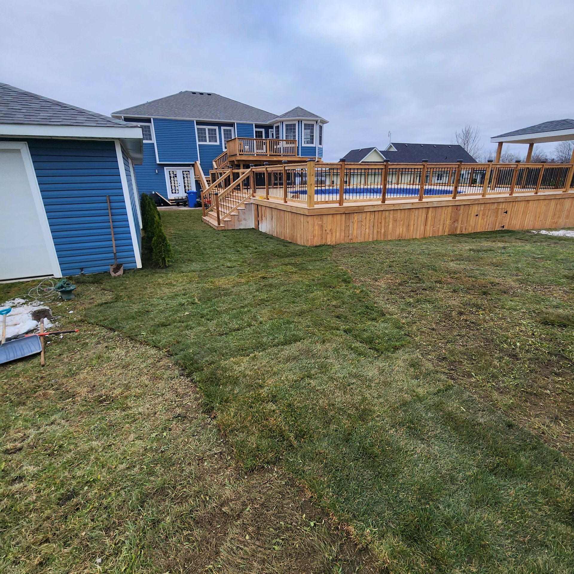 Backyard with a blue house, wooden deck, swimming pool, and shed; overcast sky.