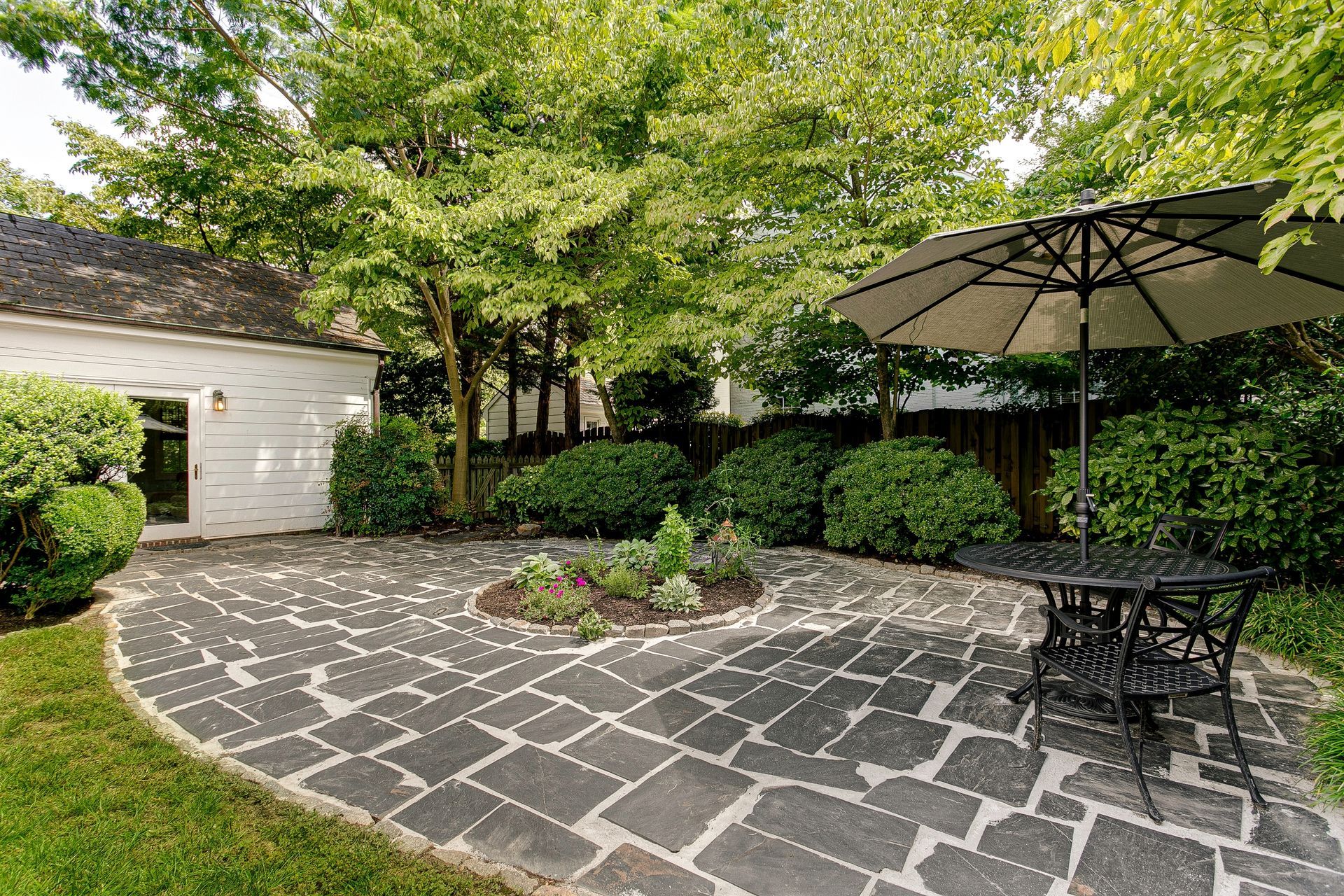 A stone patio with a table and umbrella sits in a green backyard next to a white house with trees in the background.