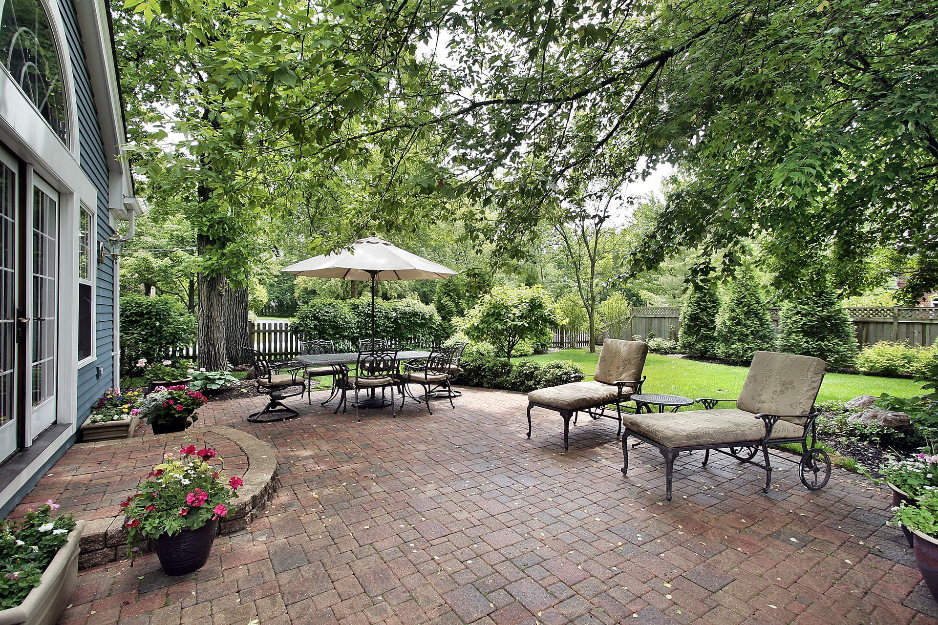 A brick patio with a dining table under an umbrella and two lounge chairs, surrounded by lush trees and greenery.