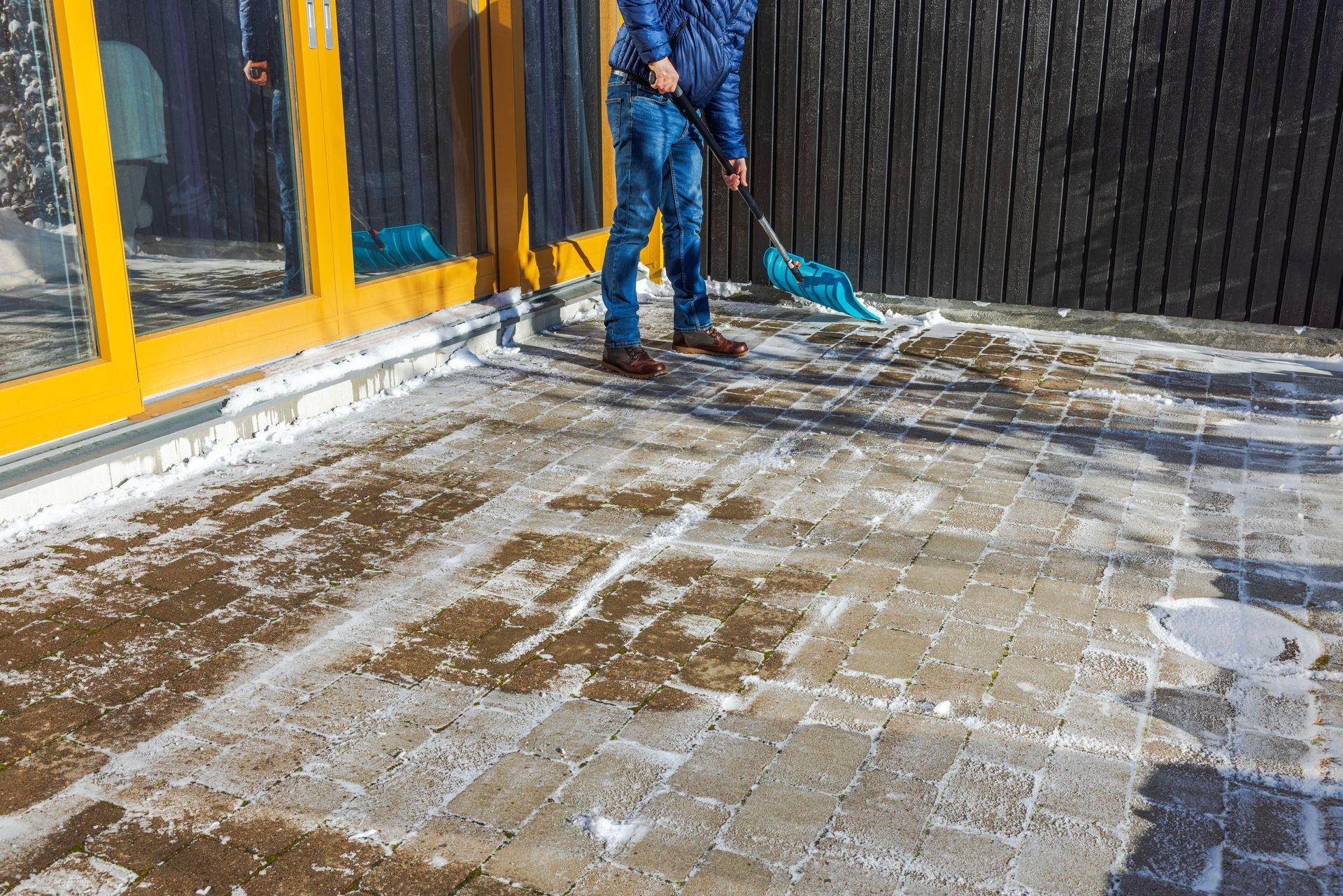 Person shoveling snow from a brick patio next to a building with yellow trim and a wooden fence.