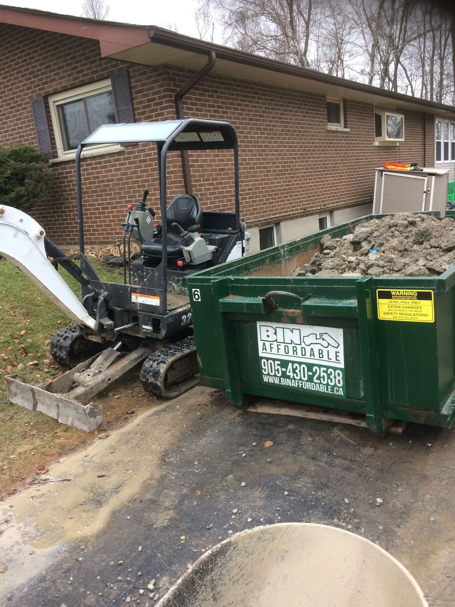 A small excavator is parked next to a dumpster in front of a brick house.